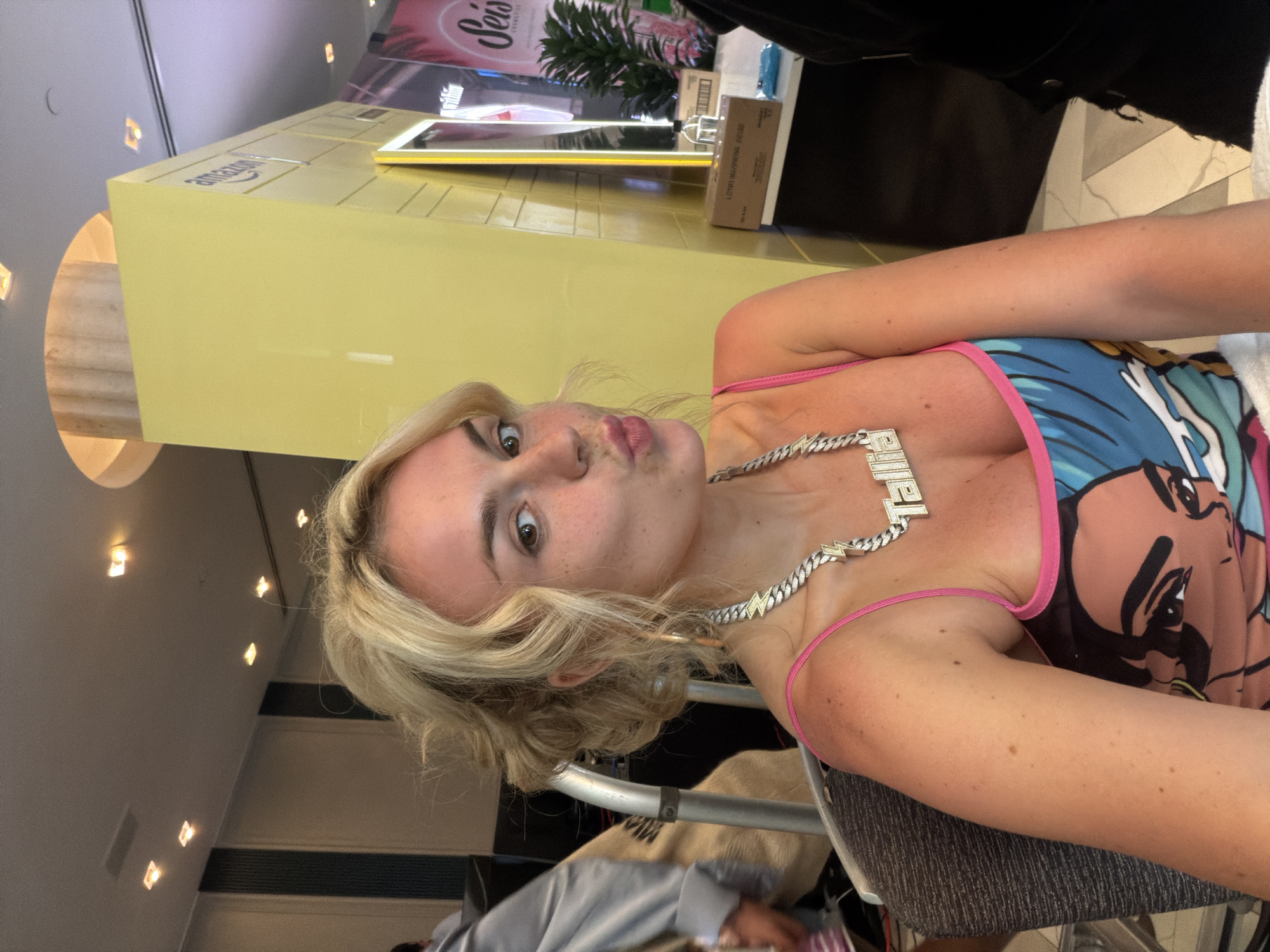 A woman with blonde hair and a chain with letters "PRIME" is looking at the camera with pursed lips, possibly taking a selfie. She is wearing a tank top with a colorful graphic design and pink straps. The background appears to be a public space, possibly a restaurant or store, with yellow fixtures and recessed lighting. An "amazon" box and plants are seen in the background. The location is likely within the city of Miami Beach, Florida, based on the presence of "amazon" and "Prime" and style of the establishment. Other people, including those wearing casual clothing, are visible in the background.