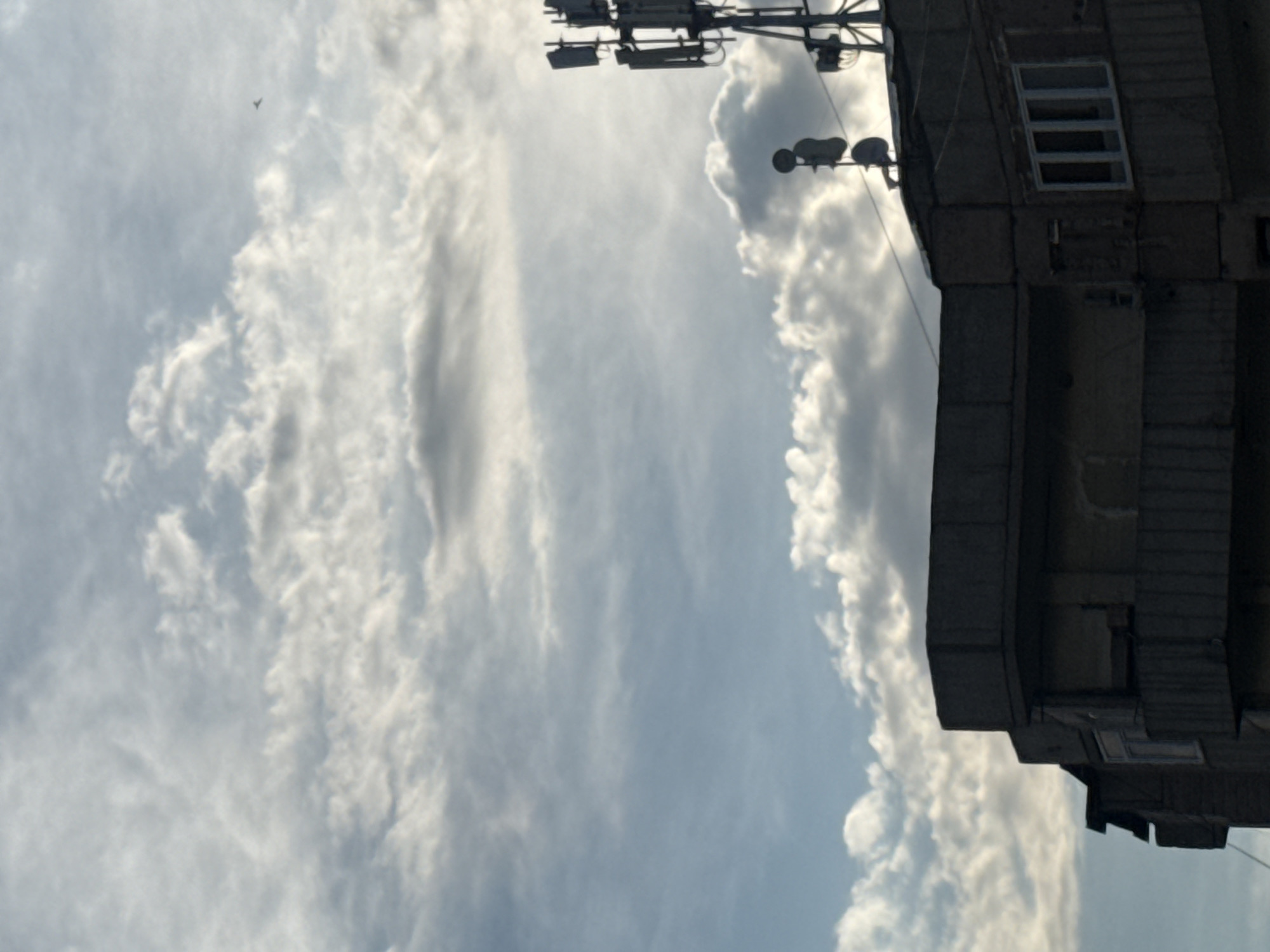 The image shows the upper portion of a concrete building against a cloudy sky. The building has balconies with railing and multiple windows. On the roof of the building, there are communication antennas and dishes. In the distance, a small object, possibly a bird or a drone, is visible in the sky. The lighting suggests it is either early morning or late afternoon. The scene is consistent with an urban environment, potentially in Abovyan, Armenia, given the architectural style and the presence of communication infrastructure commonly found in cities.
