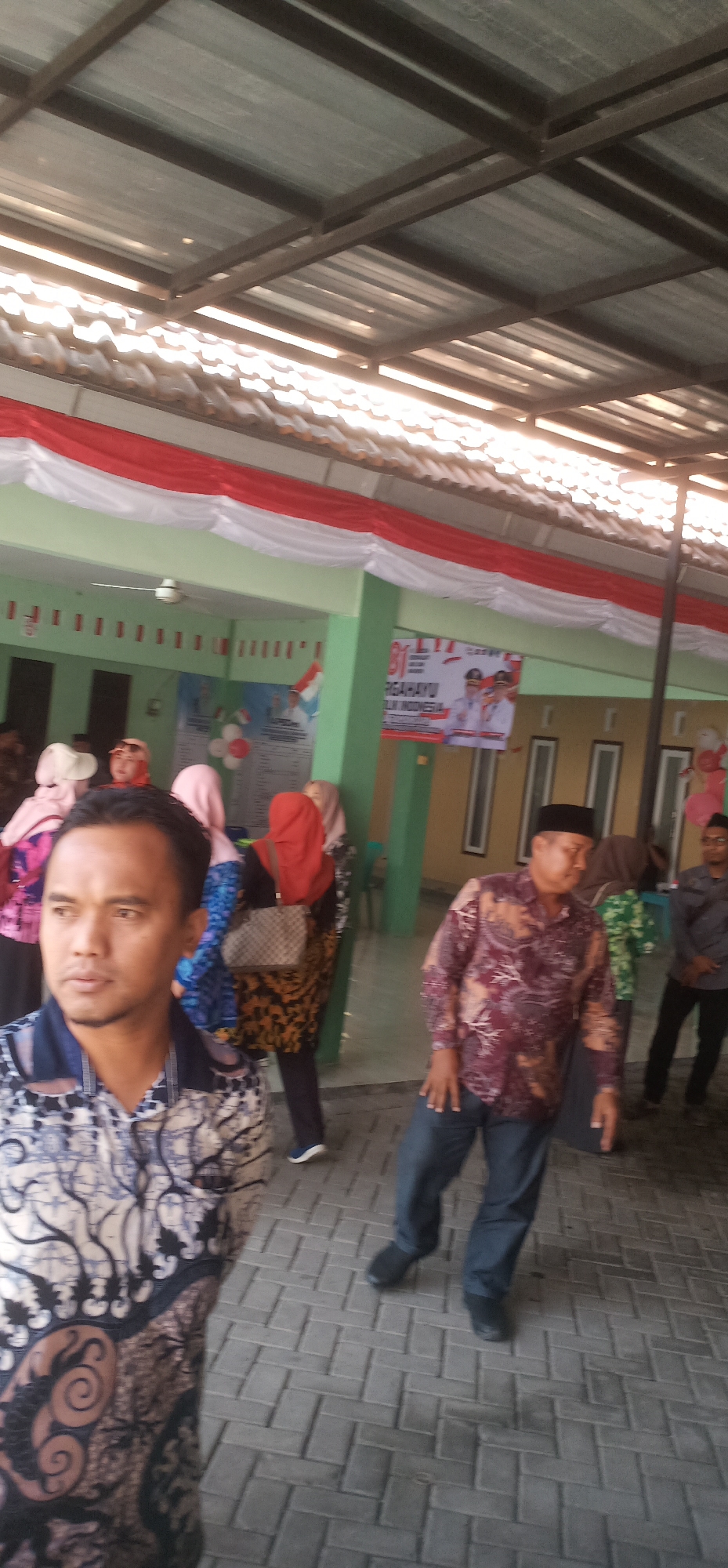 The image shows an outdoor scene, possibly a public gathering or event, at the Tamansari City of Indonesia. A corrugated metal roof supported by metal beams is visible overhead, casting shadows on the scene below. Red and white fabric banners are draped across the top of a green building in the background, decorated with national flags. Several people are present, including men and women wearing a variety of traditional and casual clothing.
In the foreground, a man in a patterned blue and white batik shirt is looking to his left. To his right, another man in a maroon patterned shirt and dark pants is walking. Other individuals are visible further back, some wearing headscarves. The ground is paved with interlocking grey bricks. A large banner with text in Indonesian and images of politicians is displayed on the green building. Balloons are also visible, suggesting a celebratory atmosphere. The overall impression is one of a community event taking place in a semi-covered outdoor area.