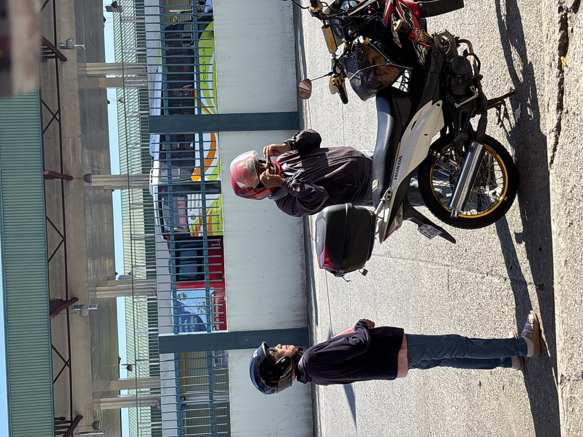 Two individuals are standing beside a parked motorcycle on a paved street, with a covered walkway and a bus in the background. The individual on the left is wearing a black helmet, a dark blue jacket, and blue jeans, with their hands clasped in front of them. The individual on the right, wearing a red helmet, is adjusting their sunglasses. The motorcycle is a white and red Honda model, with a black storage box mounted on the rear. The bus in the background is green and yellow, with "BACHELOR TOURS" written on its side. The scene appears to be located at the Tagum City Integrated Bus Terminal, Philippines, characterized by the presence of buses, a covered waiting area, and its urban setting. The lighting suggests it is daytime.