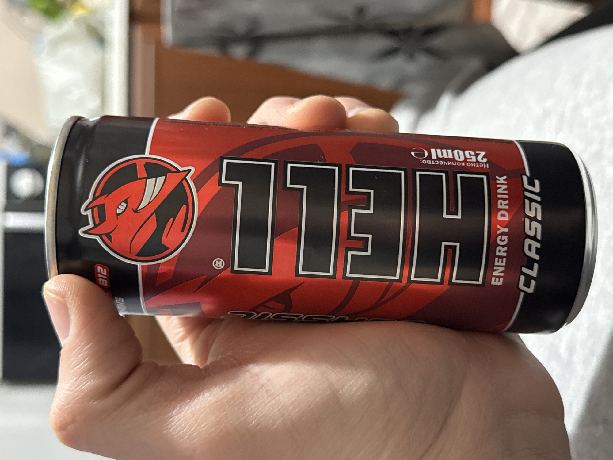 A hand holds a can of "Hell Classic" energy drink. The can is red and black with a devil logo and the word "HELL" printed on it in large, bold letters. "Energy Drink Classic" and "250ml" are also printed on the can. The background is blurred, but appears to be a room with furniture. It is impossible to determine if the image was taken in Stara Zagora, Bulgaria.