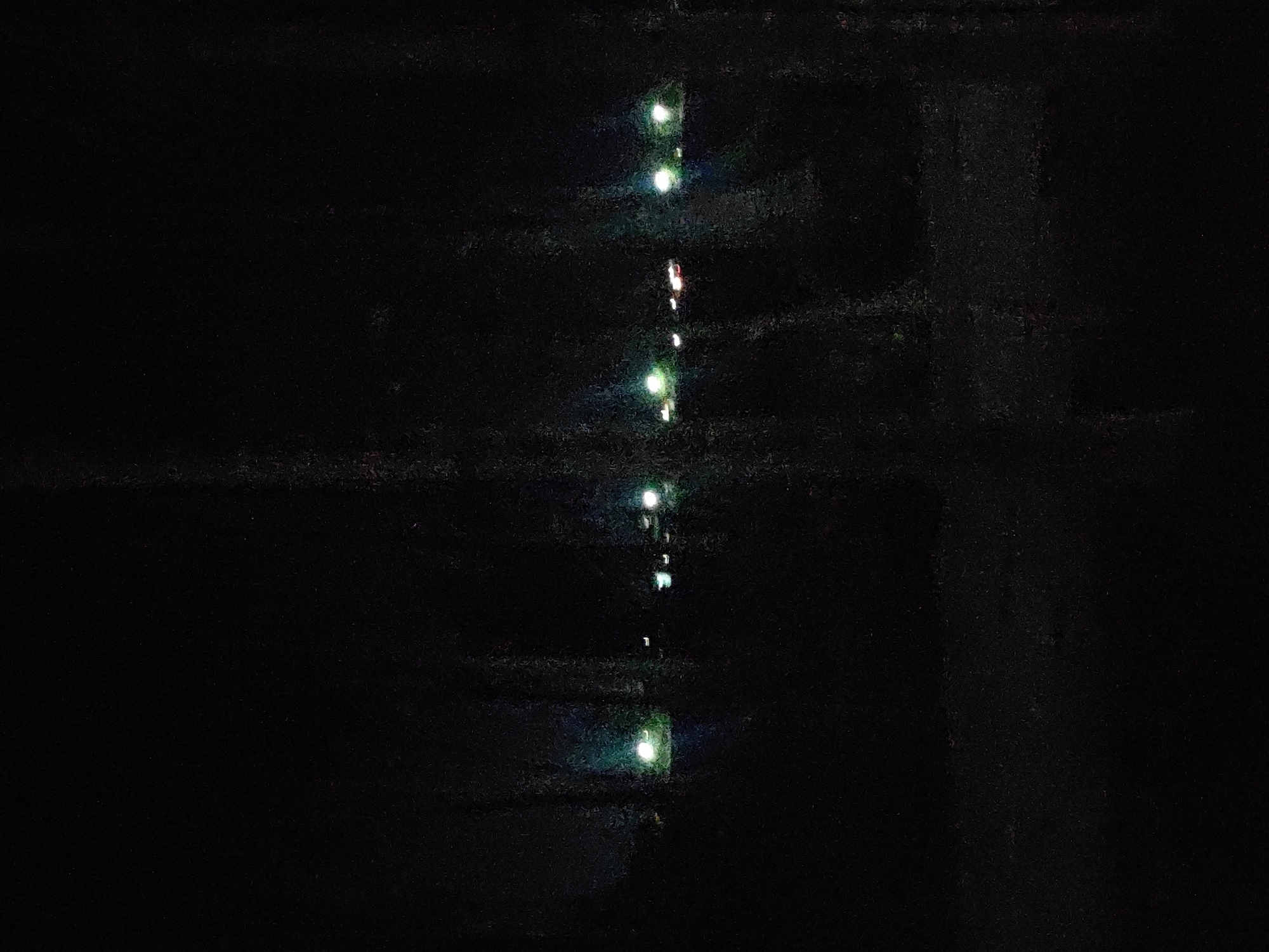 The image depicts a dark, outdoor scene with several points of light in the distance, suggesting artificial illumination from streetlights or vehicles. The foreground is dominated by deep shadows, with vertical streaks or textures that could indicate foliage, branches, or even a patterned surface like a window screen or curtain. The lighting conditions are low, and the overall impression is one of a nighttime view, possibly from indoors looking out. The specific location in Meunasahkulam, Indonesia, is not discernible from the visual information provided in the image itself.