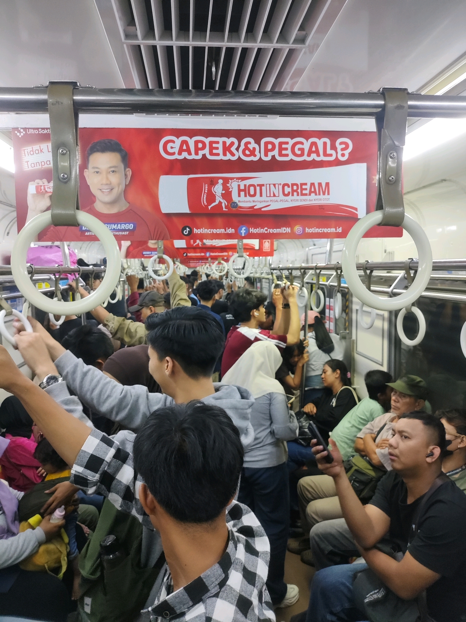 The image shows an advertisement for "Hotin Cream" displayed inside a crowded commuter train in Bekasi, Indonesia. The advertisement features a smiling man and text that reads "CAPEKI. & PEGAL?". The train car is filled with passengers standing and holding onto handrails, indicating a busy commute. The environment appears to be typical of public transportation in a densely populated urban area.