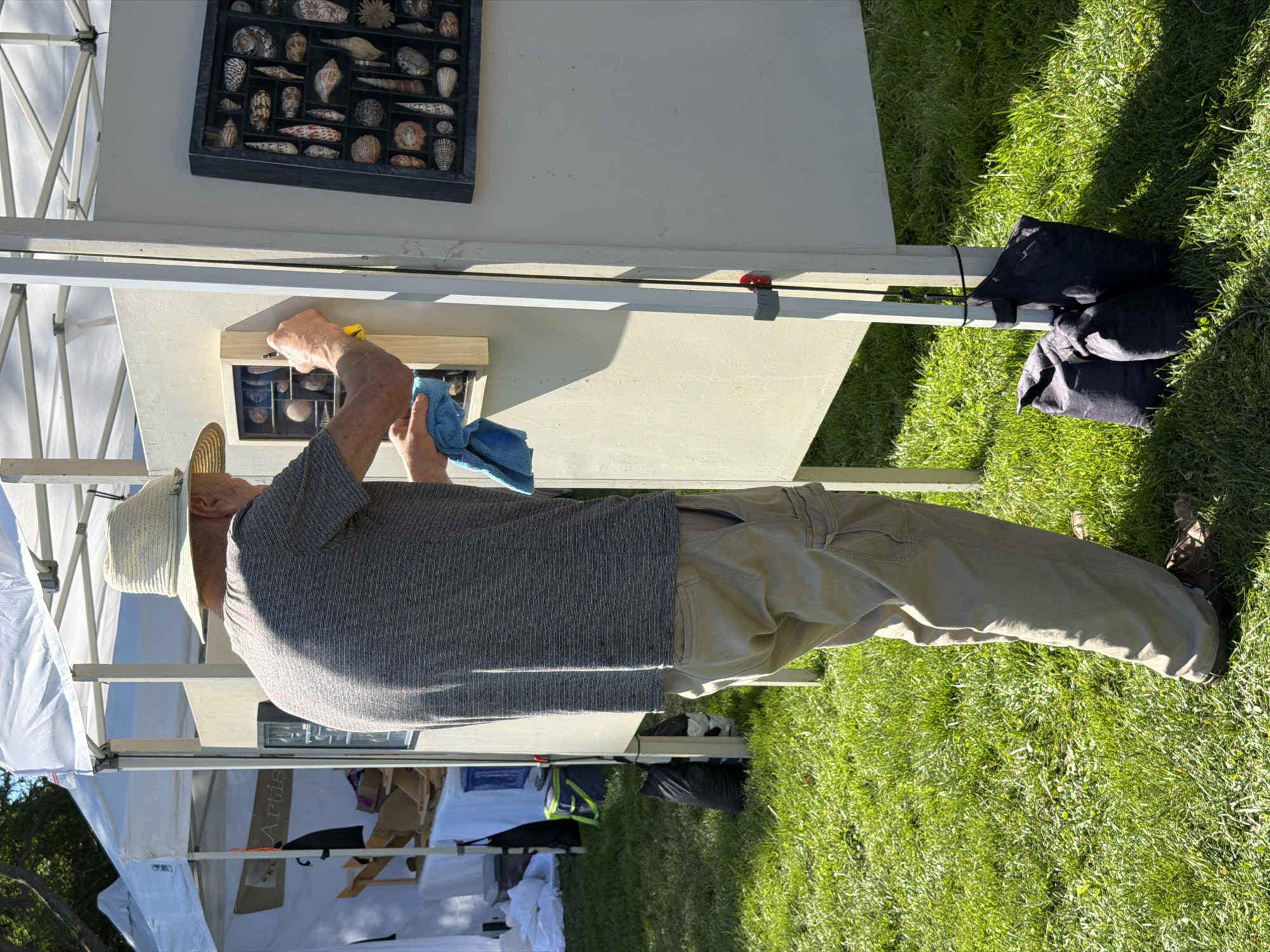An elderly Caucasian male wearing a straw hat, a striped grey t-shirt, and khaki cargo pants is cleaning a display board at an outdoor market. He is holding a yellow scraper in his right hand and a blue cloth in his left hand, and appears to be wiping the display board. The man is standing on a grassy area with a white tent structure and another display board featuring seashells to his right. The display board he is cleaning has small compartments containing shells. In the background, other tents and display items are visible. The scene is set outdoors, illuminated by sunlight. This appears to be an art or craft fair in the Village of Sag Harbor, USA.