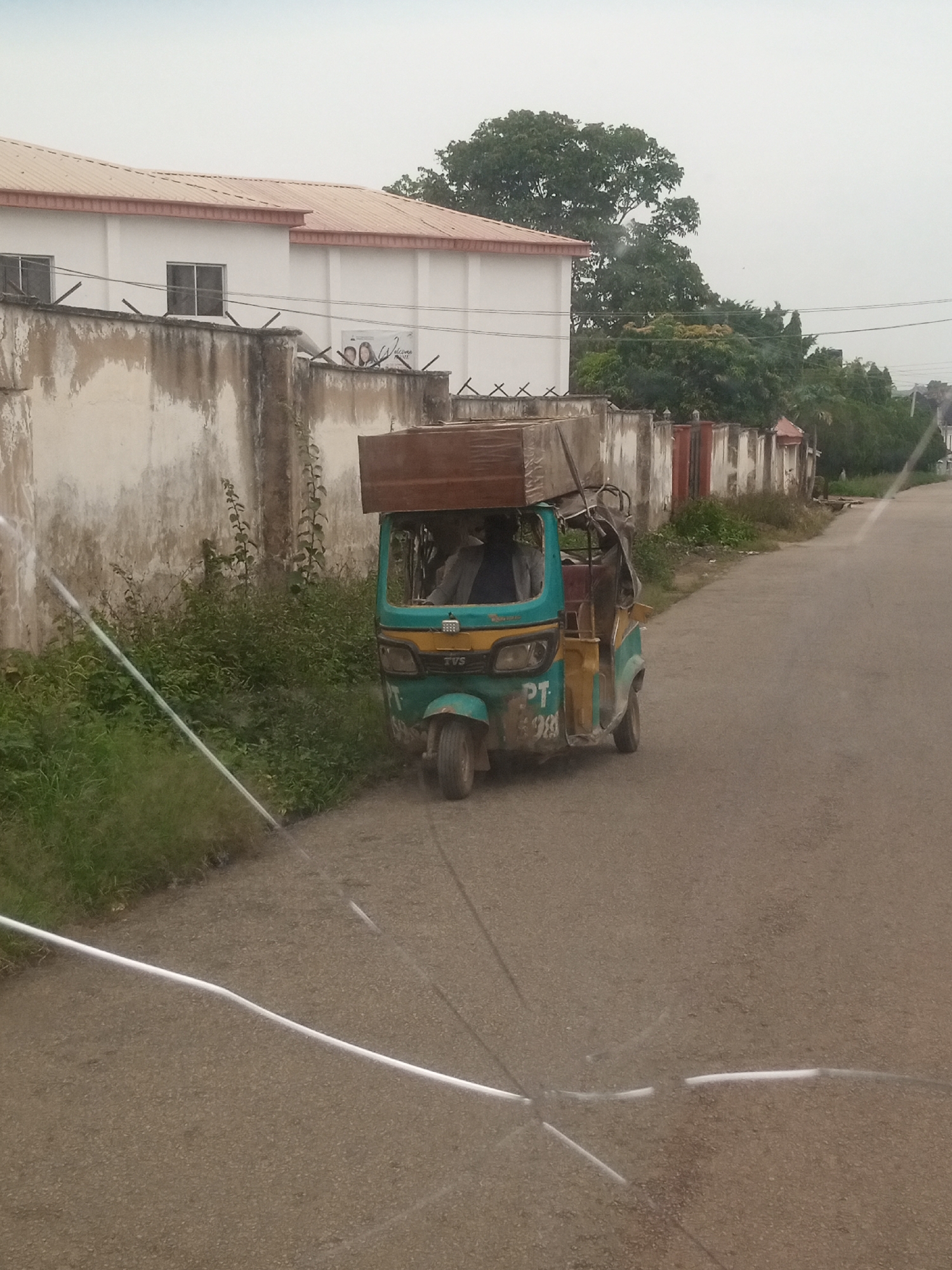 An outdoor medium shot captures a three-wheeled auto-rickshaw driving on a paved road. The auto-rickshaw, primarily teal with yellow accents, carries a large, flat, wooden object on its roof. The auto-rickshaw's driver is visible behind the steering wheel. To the left of the road, a concrete wall, weathered and stained, runs alongside a patch of lush green vegetation. Beyond the wall, a white building with a reddish-brown roof and dark-framed windows is visible. In the background, several large trees are sparsely scattered against an overcast sky. The scene appears to be in a semi-urban or suburban area.