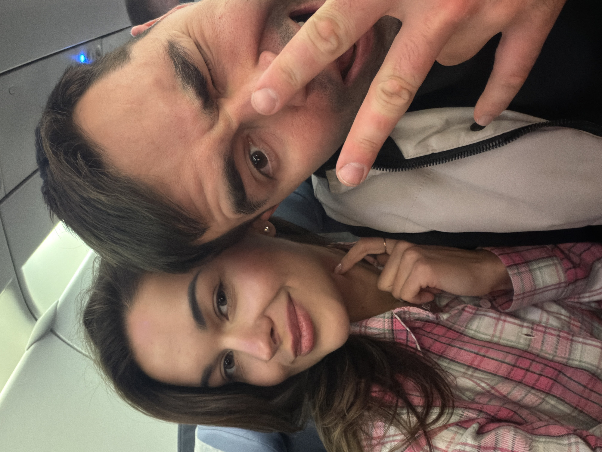 The image shows a selfie taken by a man and a woman inside an airplane. The man is positioned above the woman, and both are looking at the camera. The man is making a "peace" sign with his fingers, winking, and sticking out his tongue. He is wearing a white jacket over a dark shirt. The woman is smiling and wearing a pink and white plaid shirt. They are seated on an airplane. The location is likely in the vicinity of Емельяново (аэропорт), Russia.