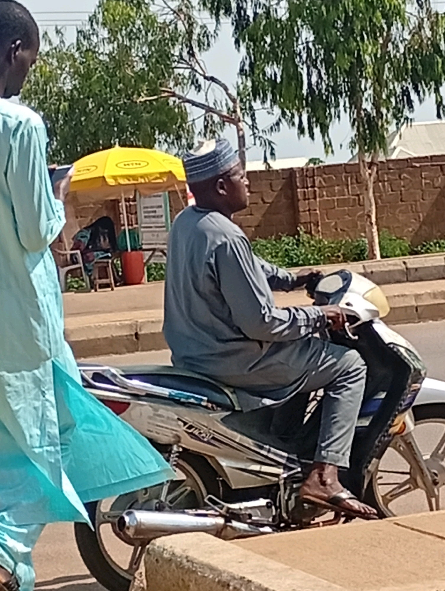A man wearing a grey tunic and trousers, and a patterned cap, is riding a white and silver motorcycle. He is looking to the left. Next to him, on the left, a person in a light blue garment, possibly a robe, is walking away from the camera. In the background, a yellow umbrella with the "MTN" logo is open over a stall. Behind the stall, a brick wall is visible. The scene appears to be in an outdoor urban environment, possibly in Tumfure, Nigeria.