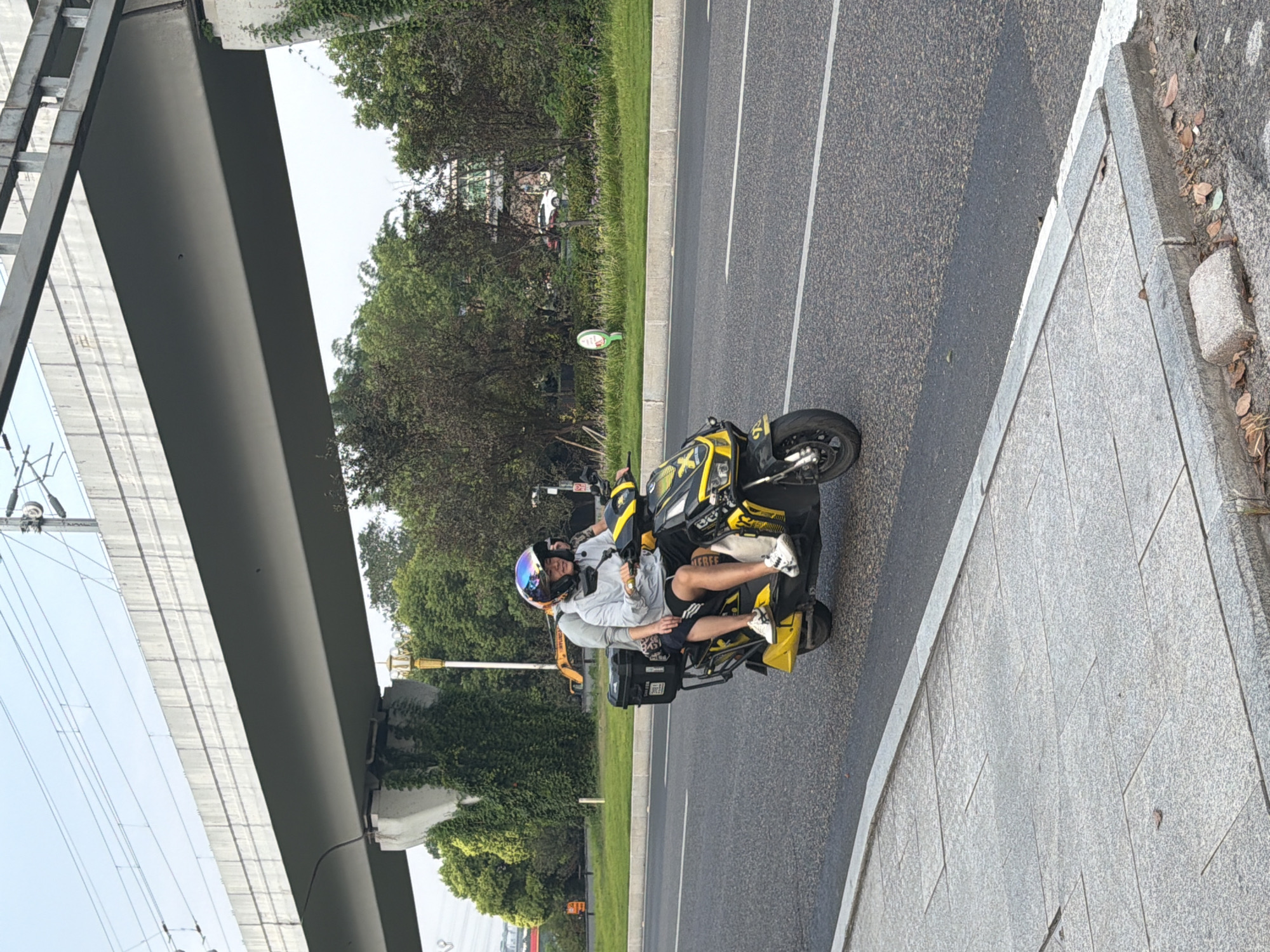 In the city of Jiangjin, China, two individuals are riding on a yellow and black electric scooter. The driver, wearing a helmet with a rainbow-colored visor and a grey long-sleeved shirt, operates the scooter. Behind the driver sits a passenger, also wearing a helmet. The scooter is equipped with a black storage box on the rear. They are traveling on a paved road, with a sidewalk to the left and a grassy area with trees and a bridge overhead to the right. The sky is overcast.