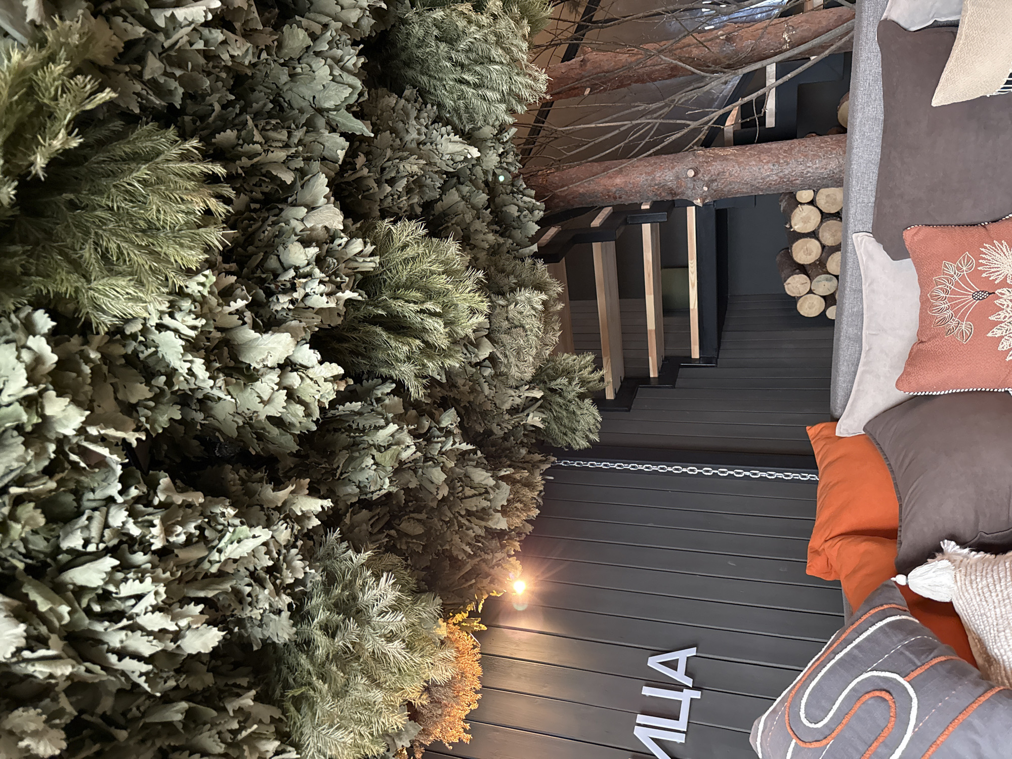 The image captures an interior scene in Vyakhtelevo, Russia. A section of a building is visible, including a wall made of horizontal wooden planks, along which the Cyrillic word "ВИЛЛА" (villa) is written in white lettering. Hanging from a chain is a dried plant arrangement, possibly foliage. The interior decor includes large tree branches and logs used as structural or decorative elements. Nearby, a pile of logs is stacked for possible heating or decorative purpose. Adjacent to the wall is a section of a lounge area with several pillows in varying colors and textures, including white, gray, orange, and a brown pillow with a white floral design.