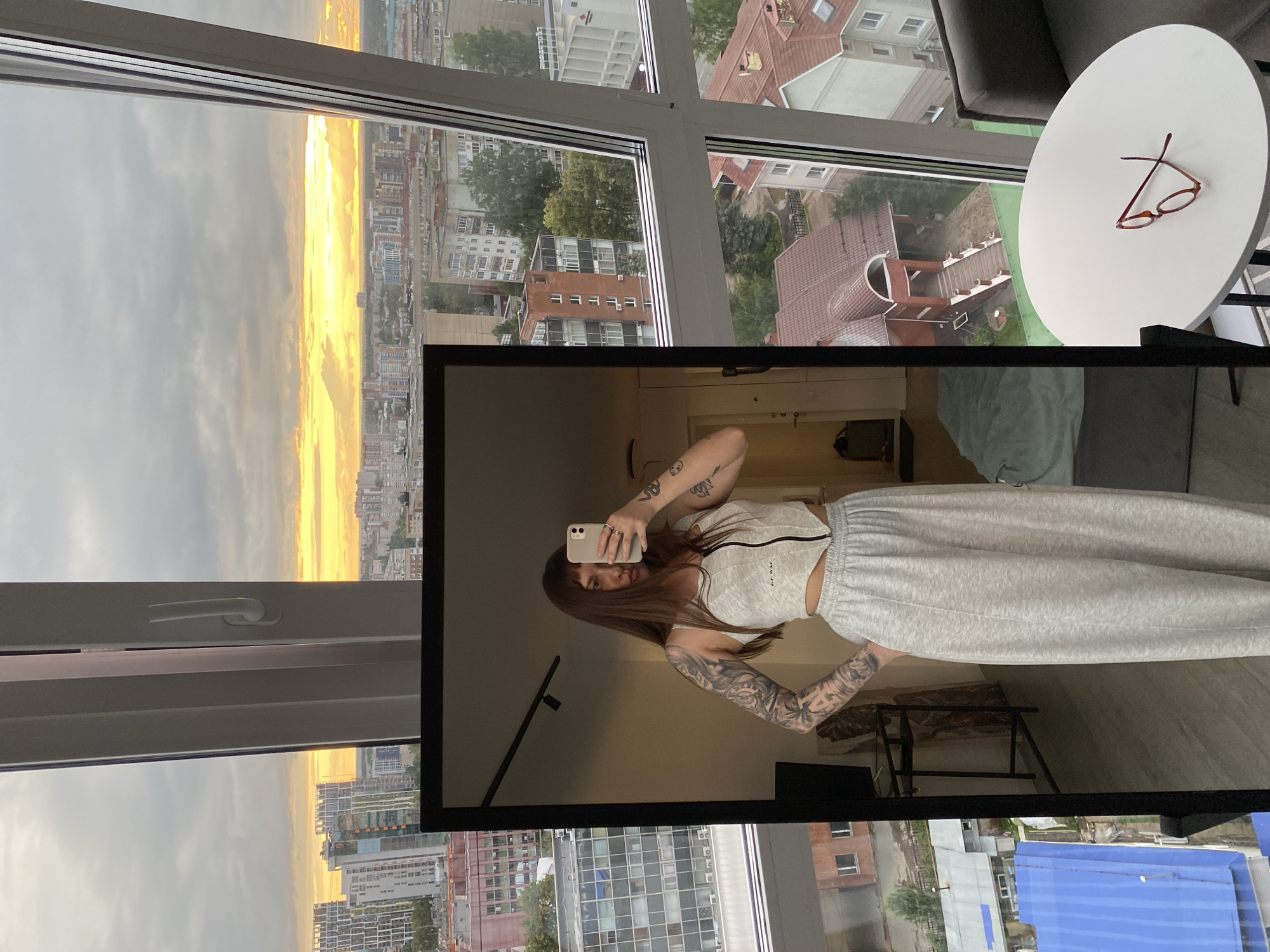 The image shows a young woman with long brown hair and extensive tattoos on her arms, taking a selfie in a full-length mirror. She is wearing a light gray crop top with a zipper and matching wide-leg sweatpants. The mirror reflects her and a portion of the room behind her, including a bed on the floor and a tall cabinet. Outside the large windows, a cityscape is visible, with numerous apartment buildings and houses. The sky is cloudy with a bright orange and yellow sunset breaking through. The scene is set in Izhevsk, Russia, likely in a modern apartment building with a panoramic view of the city. On a round white table to the right of the frame, a pair of red-rimmed glasses rests.