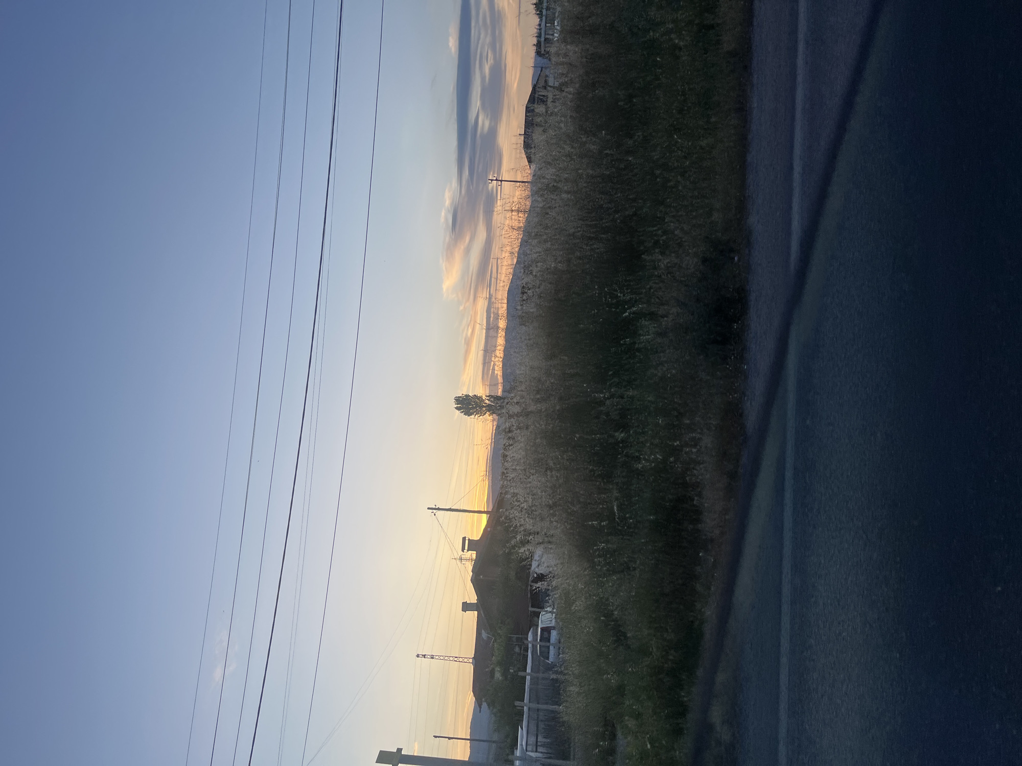 The image depicts an outdoor scene, possibly a rural area near Blagoevgrad, Bulgaria, during what appears to be the early morning or late evening based on the light and the position of the sun. The scene includes:
1. **Sky:** A clear sky with variations of light blue and hints of orange and pink near the horizon, indicating either sunrise or sunset. Streaks of clouds are visible as well.
2. **Power Lines:** Multiple parallel power lines stretch across the upper-left part of the image.
3. **Vegetation:** Tall, dense vegetation fills the right side of the frame, potentially including grasses and shrubs.
4. **Buildings/Structures:** Several buildings and structures, including what appears to be a water tower and power lines, are visible in the middle ground. Some residential structures are noted.
5. **Objects:** Other objects include what looks like a parked vehicle and a fence.
6. **Environment:** The environment is somewhat rural or suburban, with a mix of natural and built elements. The lighting suggests a quiet, peaceful atmosphere.
7. **Vehicle Interior:** There is a glimpse of what appears to be the interior of a vehicle on the right side of the picture, possibly indicating that the photo was taken from inside a car.
The location within Blagoevgrad is unidentifiable without more context.