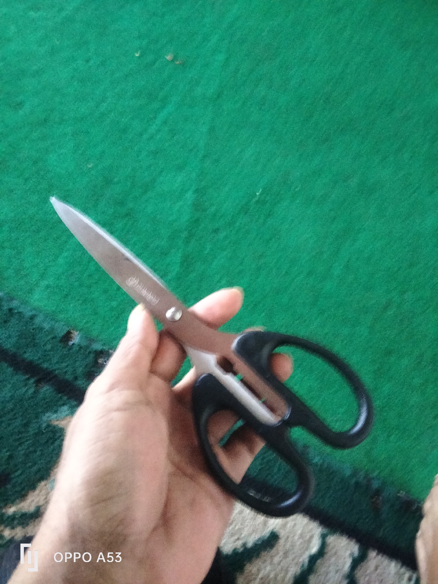 An overhead shot shows a hand holding a pair of scissors against a green carpet. The scissors have silver blades and brown and black handles. The OPPO A53 logo is visible in the bottom left corner. The setting appears to be a room in Badung, Indonesia.