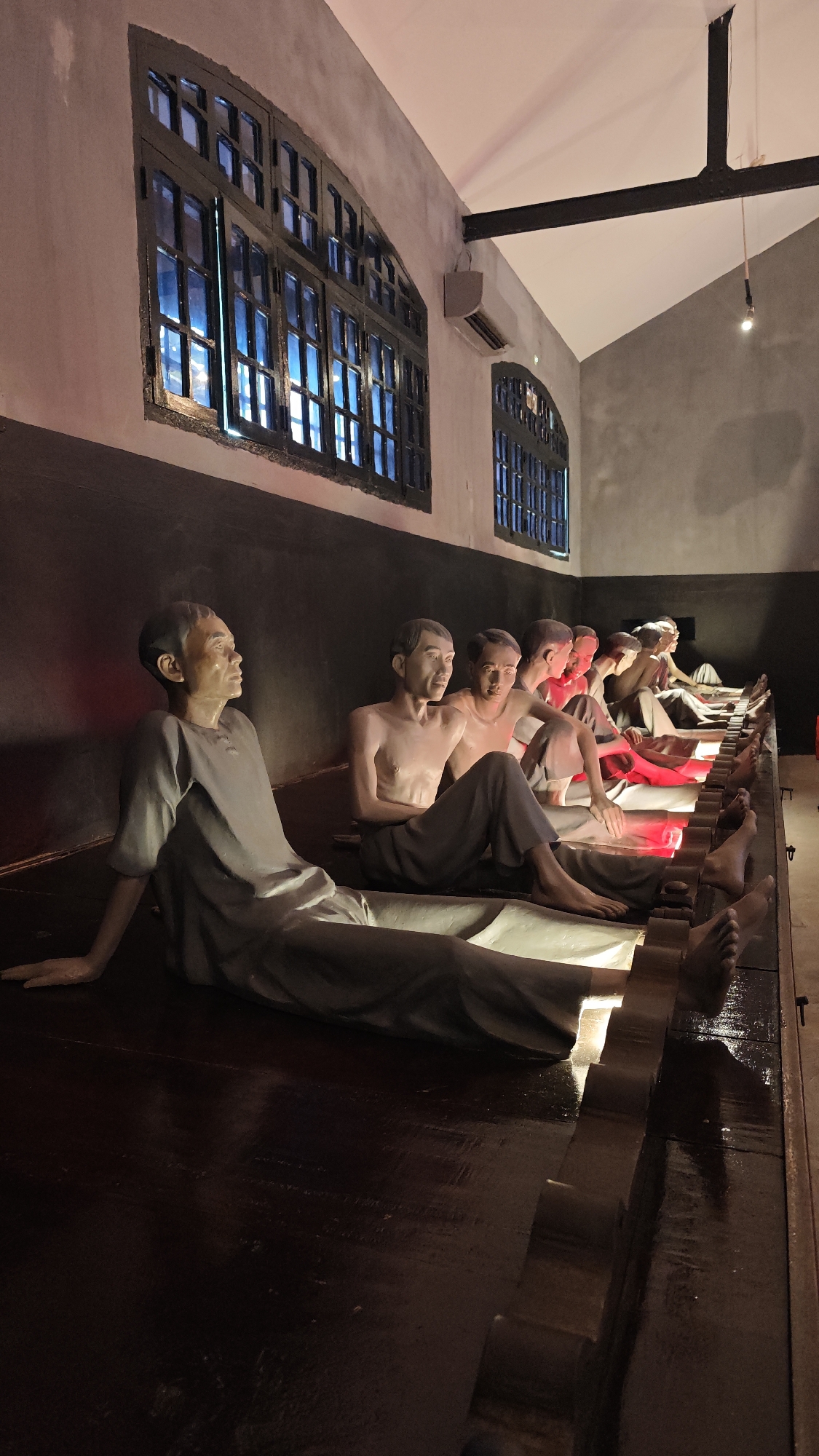 The image depicts a scene inside the Hỏa Lò Prison (also known as the Hanoi Hilton) in Hà Nội, Vietnam. The scene shows a row of wax figures depicting prisoners sitting on a raised platform. The figures are in various states of dress, some wearing prison garb and others shirtless. There are two large windows on the wall behind the figures. Red lights illuminate the figures' feet. The platform they are seated on appears to be made of dark wood. The overall environment is dimly lit.