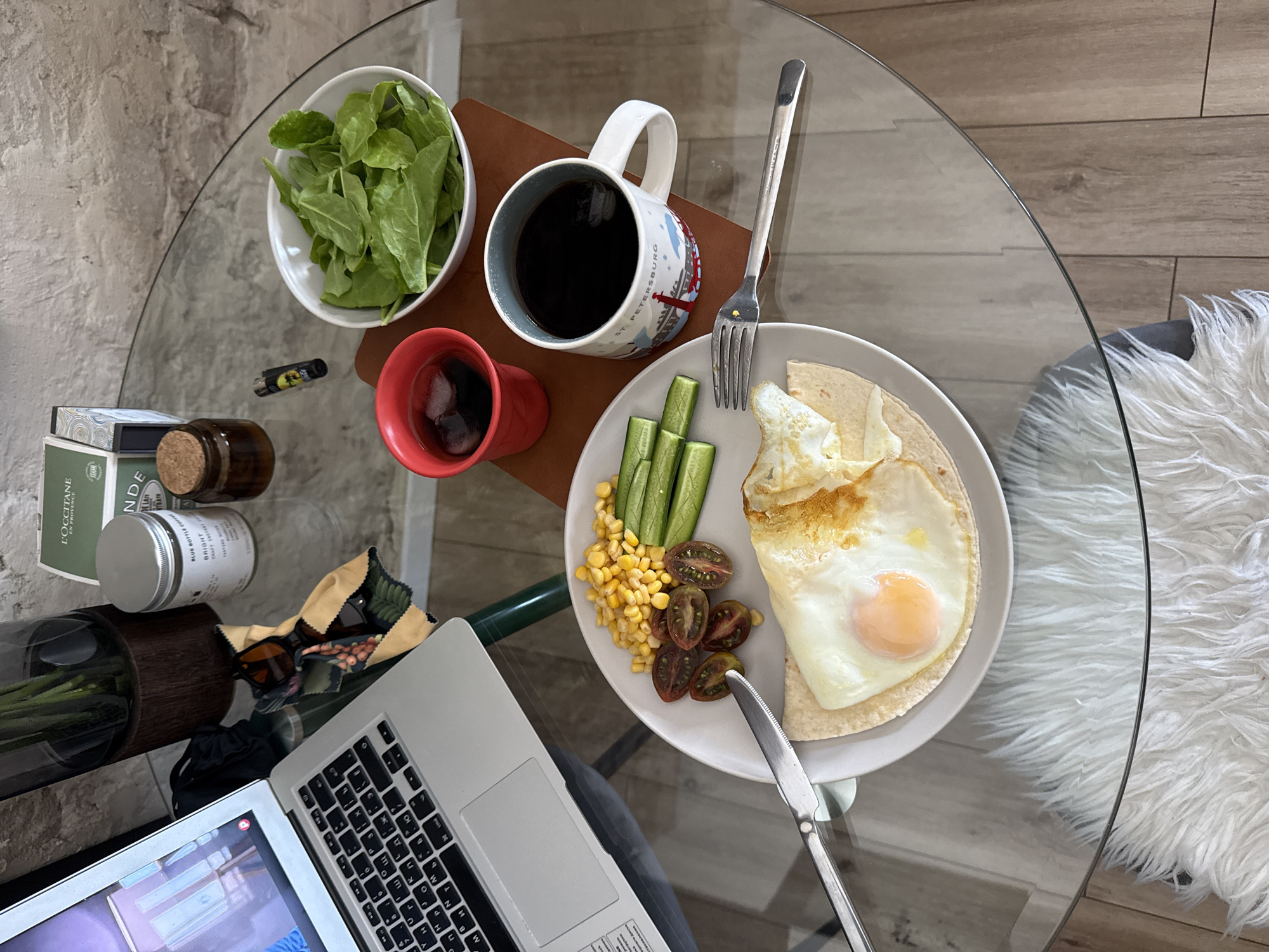 The image shows a glass-topped round table set with a variety of items suggesting a meal and work situation. On the left side of the table are several cosmetics or skincare products, a vase with green stems, and a partially visible laptop displaying a video.
Towards the center of the table, from left to right, there is a small white bowl filled with green leaves, possibly salad; a small red mug containing a dark liquid; and a mug with a design featuring the city "St. Petersburg," filled with black coffee. These items are placed on a brown rectangular tray.
A plate is positioned to the right of the coffee mug, holding a meal that includes an egg, corn, cucumber sticks, and dark cherry tomatoes arranged on a tortilla. A fork and a knife are set beside the plate.
The table is placed on a surface with a light wooden pattern, and a white fluffy material, potentially a rug or throw, is visible on the right side of the table. The scene appears to be indoors, and the lighting is bright. The image does not suggest the city of Tel-Aviv, Israel or any specific location.