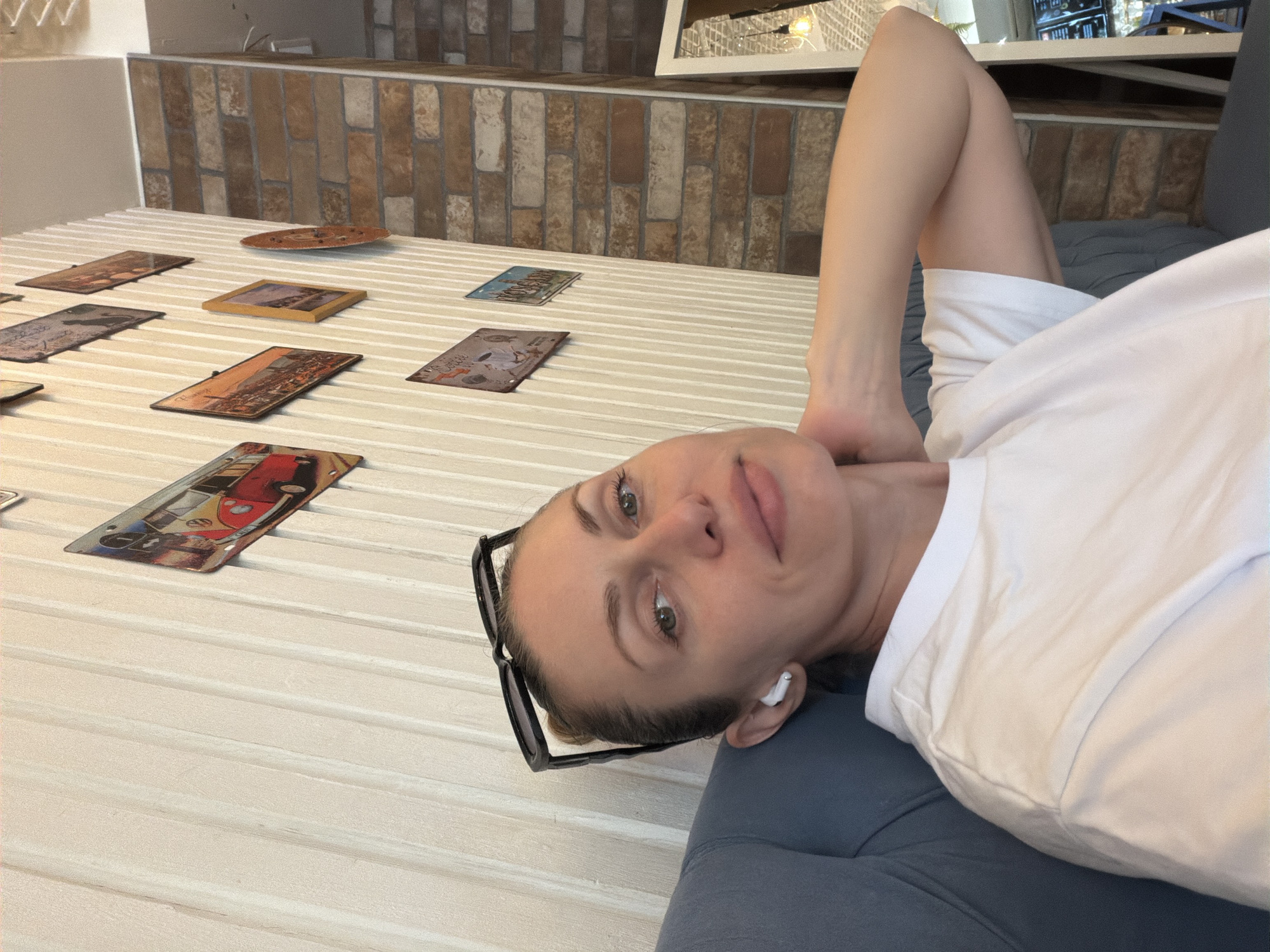 The media file features a woman lying on her side, looking at the camera. She is wearing a white t-shirt and glasses, and has earbuds in her ears. Her head is tilted, and she appears relaxed. The background shows a wall with a brick pattern and a ribbed surface. Several small, rectangular objects with images on them are scattered across the ribbed surface. One of these objects depicts a red and yellow camper van. The lighting suggests it is daytime. The setting appears to be indoors, possibly a cafe or a room with decorative elements. The woman's expression is calm and pleasant. No legible text is visible on the objects in the image, except for some faint lettering on one of them that is too small to read. The scene is static, with the woman posing for the photo.