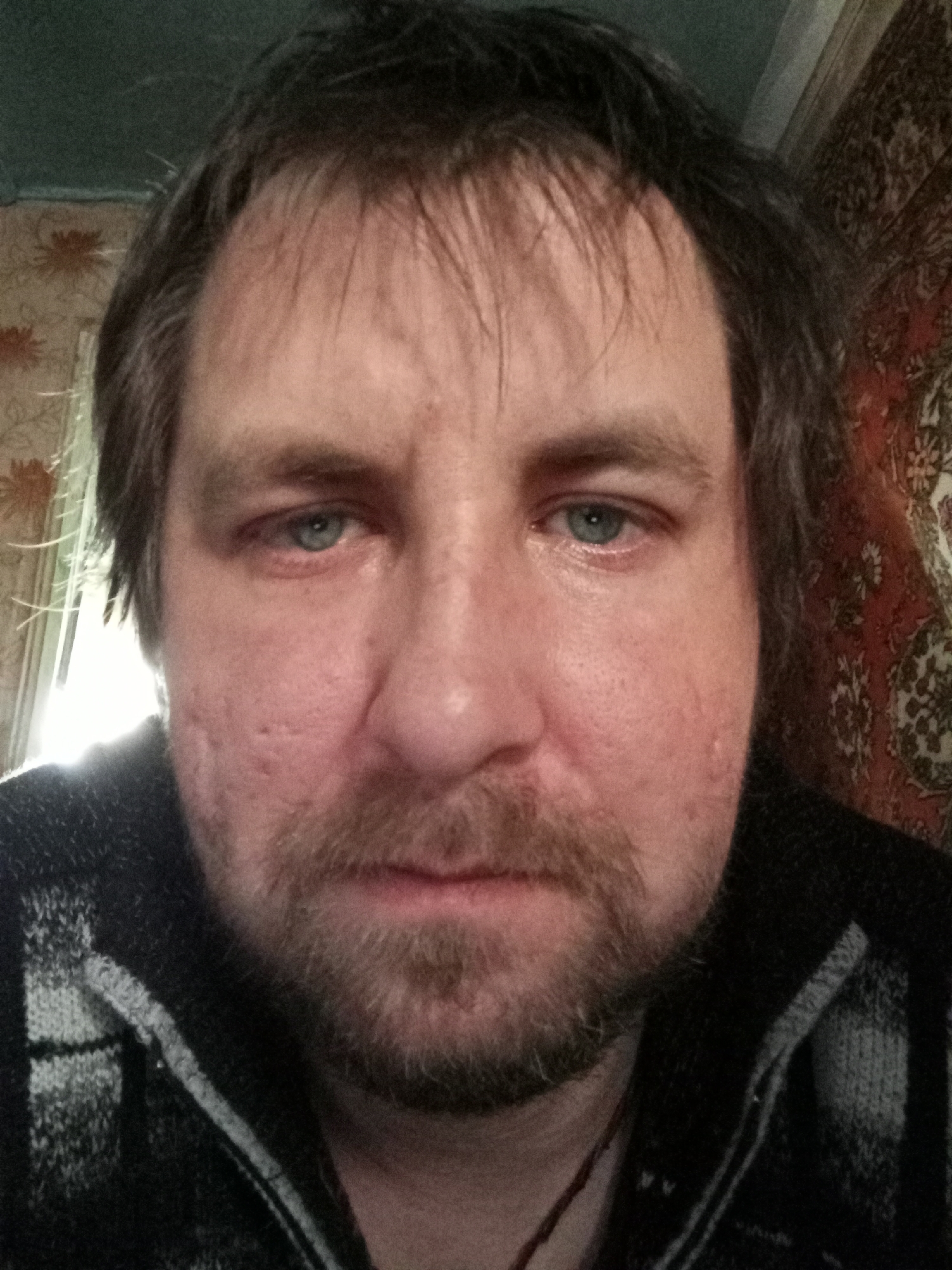 A close-up selfie of a middle-aged man with light blue eyes, short brown hair, and a beard. He appears to be wearing a dark-colored, knit sweater or jacket with a light gray stripe. The background includes a wall with floral wallpaper and a window with light coming through it. No specific location in Parabel, Russia, can be ascertained from this image.
