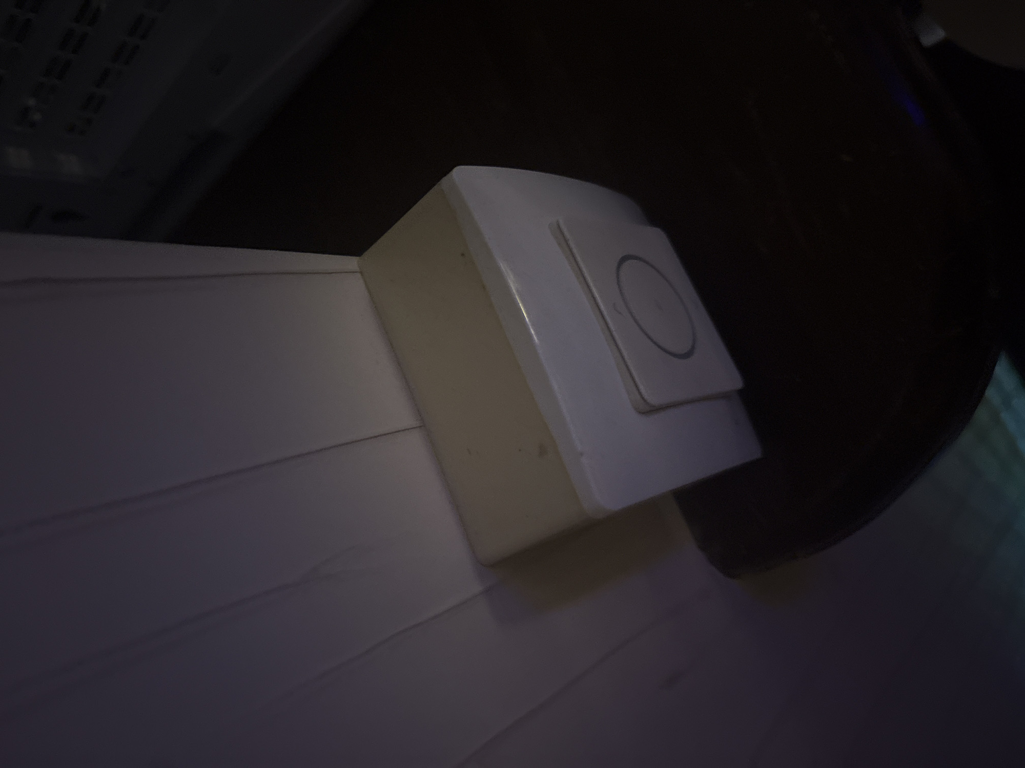 A white doorbell is mounted on a wooden wall. The doorbell is rectangular and has a circular button in the center. The wall is painted white and has vertical lines. The doorbell is located in a dimly lit room. The background is dark and out of focus. The image appears to be taken from a low angle.