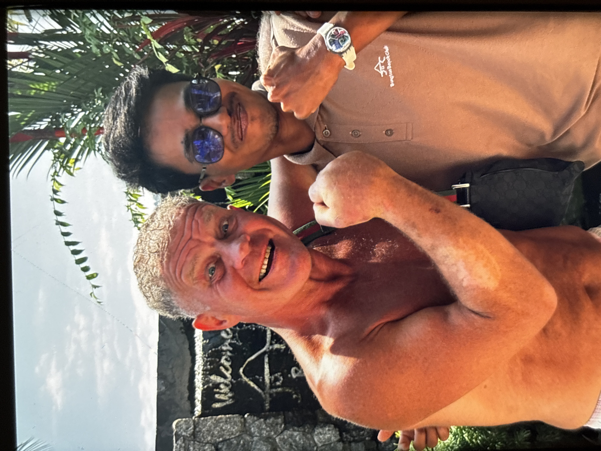 In Chalong, Thailand, two men are pictured outdoors, with a stone structure featuring the word "Welcome" visible in the background. The man on the left, who appears to be Caucasian with fair skin, is shirtless and has light-colored hair. He is smiling with his teeth showing. The man on the right, who appears to be of South Asian descent, is wearing a tan polo shirt and sunglasses. Both men are making fists with their hands. The man on the right has a watch on his left wrist that reads "Bangtao Beach Club". Lush green foliage and red-leafed plants surround the individuals, and a bright sky with scattered clouds is visible overhead.