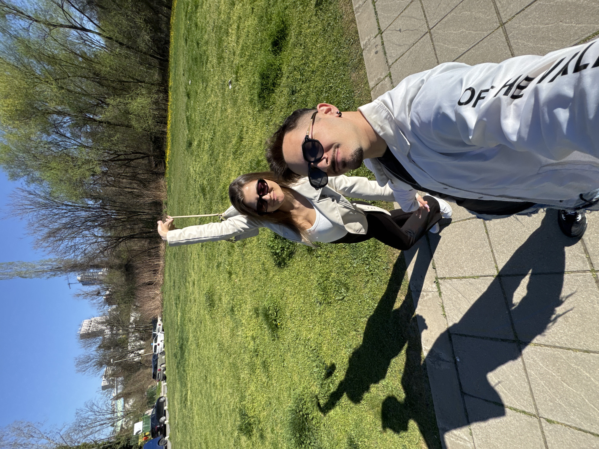 A selfie was taken by a man and woman in a park on a sunny day. The man is on the right wearing sunglasses and a white jacket with writing. The woman is on the left wearing sunglasses and a light-colored jacket over a white top. They are standing on a paved surface next to a large grassy area. Trees are in the background, and buildings are visible in the distance under a blue sky. Cars are parked along the edge of the park. The location is likely in Sofia, Bulgaria.