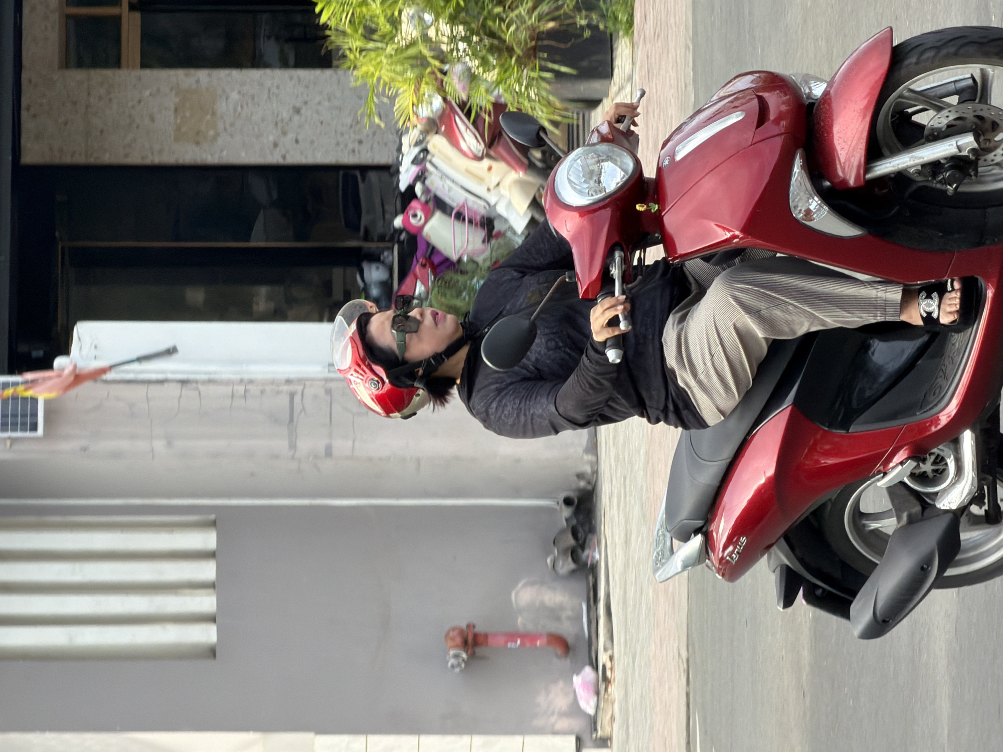 A woman wearing a black long-sleeved shirt, grey striped pants, black sunglasses, and a red and white helmet rides a red scooter. She is wearing black sandals with a silver logo. The scooter has a chrome rear-view mirror and a silver passenger grab handle. The background shows a building with a grey facade, a red fire hydrant on the left, and several other scooters parked behind her. Green foliage is visible on the right side of the image. The scene appears to be a street in Nha Trang, Vietnam.