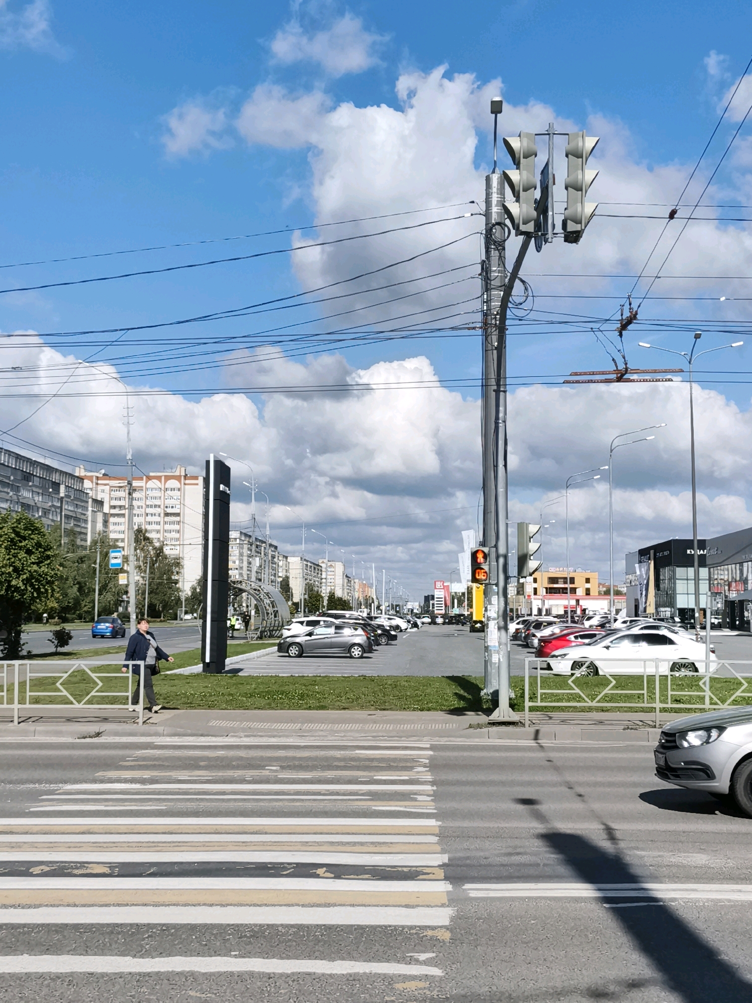 The image depicts a sunny day in Yoshkar-Ola, Russia, focusing on a street scene with a pedestrian crossing in the foreground. A woman in a dark jacket and pants walks across a pedestrian zone, which has white and yellow striped markings on the asphalt. The background reveals a city street lined with multistory apartment buildings and commercial establishments. A traffic light with an orange light illuminated is visible on the right side of the image. Several cars are parked along the street and in parking lots, including white, silver, and red vehicles. Overhead, numerous power lines crisscross the sky, partially obscuring the bright blue sky with fluffy white clouds. The overall impression is that of a typical urban environment on a clear day.