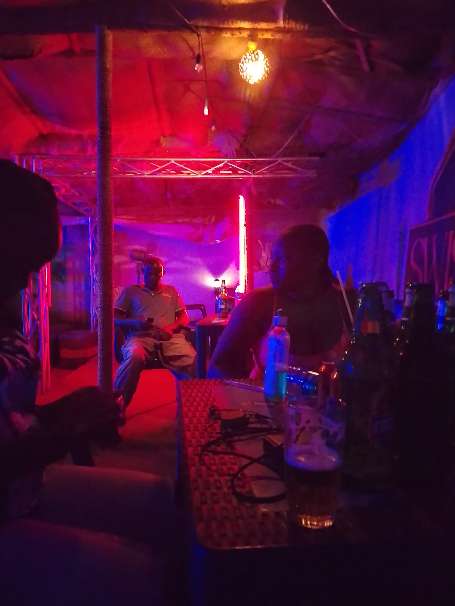 The image shows an interior scene in an unknown location in the city of Unknown, Unknown, illuminated by red and blue lighting. A man sits in a chair in the background, looking at his phone. In the foreground, there's a table with several bottles of beer and a glass of beer. Two people are seated near the table. A decorative light hangs from the ceiling.