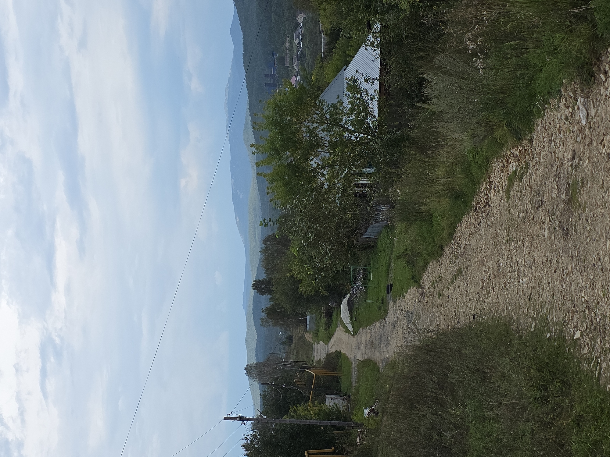 The image depicts a rural landscape with a gravel road leading through a sparsely populated area. In the foreground, the road is flanked by overgrown grassy areas and scattered debris. Several utility poles with overhead wires are visible along the left side of the road, one of which has yellow gas pipes attached. Further down the road, various structures, including houses with metal roofs and sheds, are scattered among trees and vegetation. In the background, rolling hills covered in trees recede into the distance. A large industrial complex with tall structures and smokestacks is visible on one of the distant hills, suggesting the proximity of industry to this rural setting, likely within or near the city of Magnitka, Russia, known for its industrial activities. The sky is overcast with scattered clouds. There are no people visible in the image.