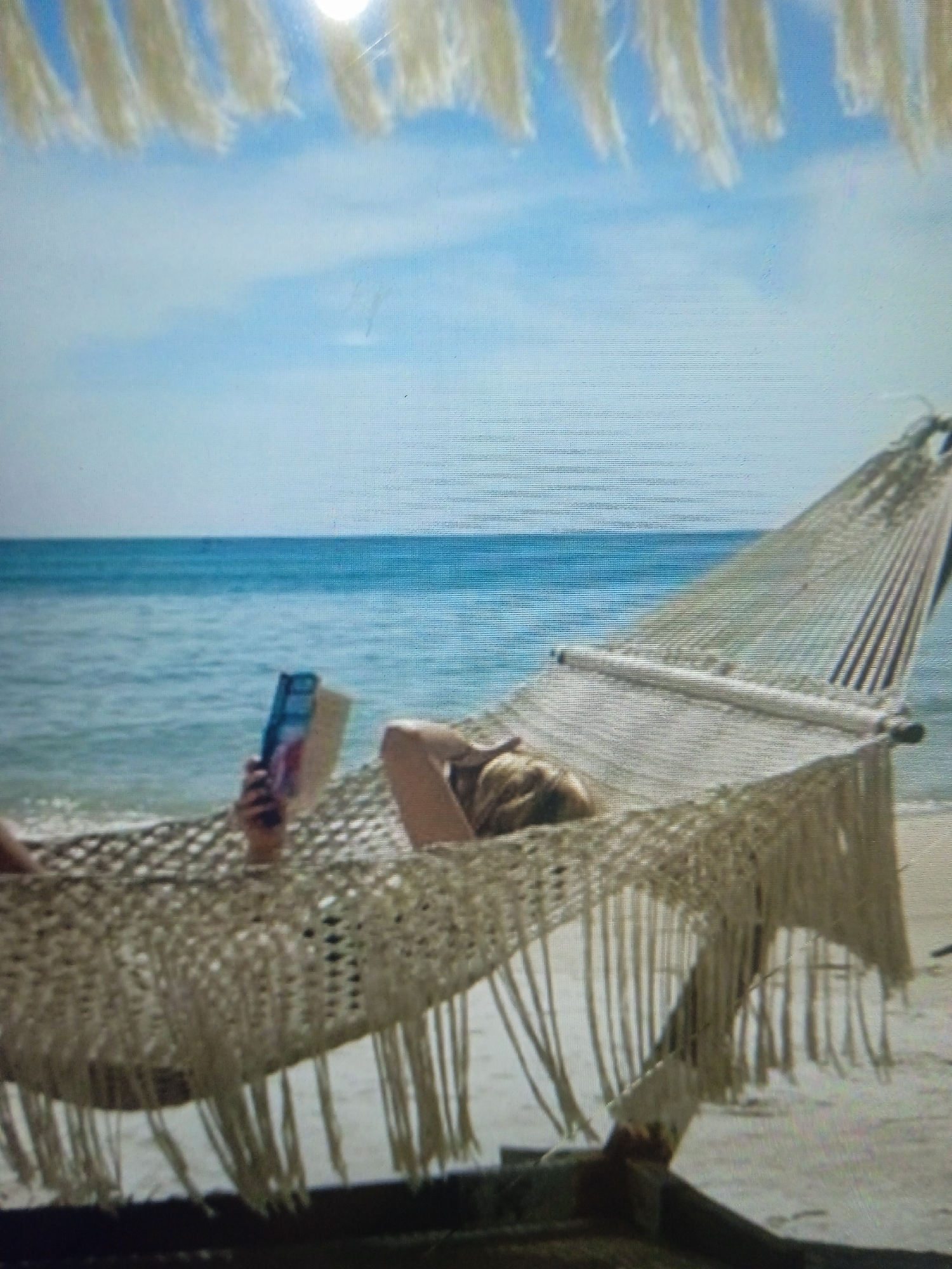 A person is lying in a hammock on a beach, reading a book. The hammock is made of rope and has a fringe of tassels. The person's face is obscured by the book, but their arm is visible. The background shows a calm blue ocean and a clear sky with some clouds. The image is taken from a slightly elevated angle, looking down at the hammock and the beach. The location is not Chernivtsi, Ukraine.