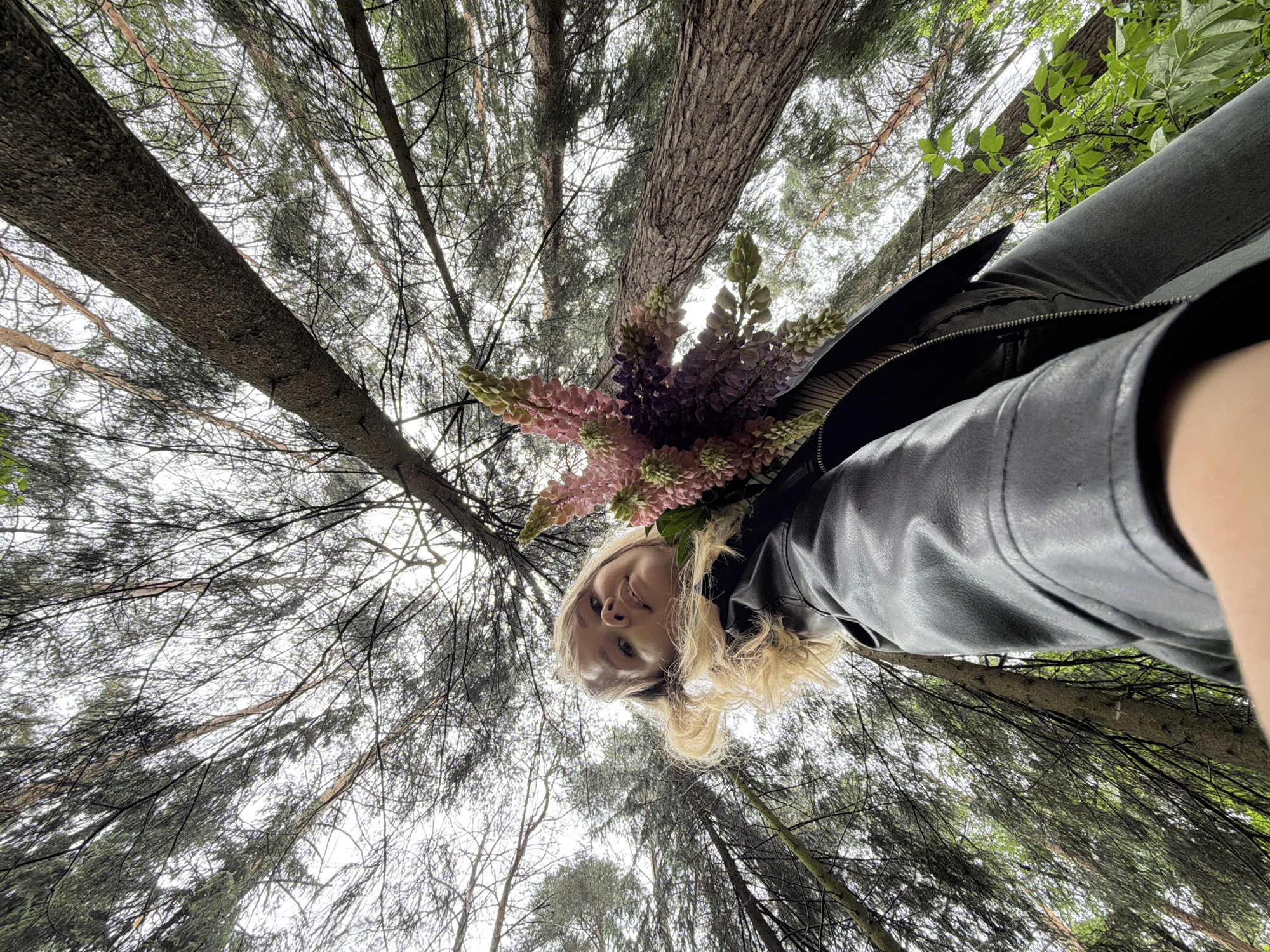 A low-angle shot looking up at a person holding flowers in a forest or park. The person, a woman with blond hair, is wearing a black leather jacket. She's holding a bouquet of lupine flowers in shades of pink and purple. The background is filled with tall pine trees reaching toward the sky, their branches and needles creating a dense canopy. The photo appears to have been taken in daylight but with overcast conditions, filtering the light through the trees. This picture was taken in the city of Chaikovsky, Russia.