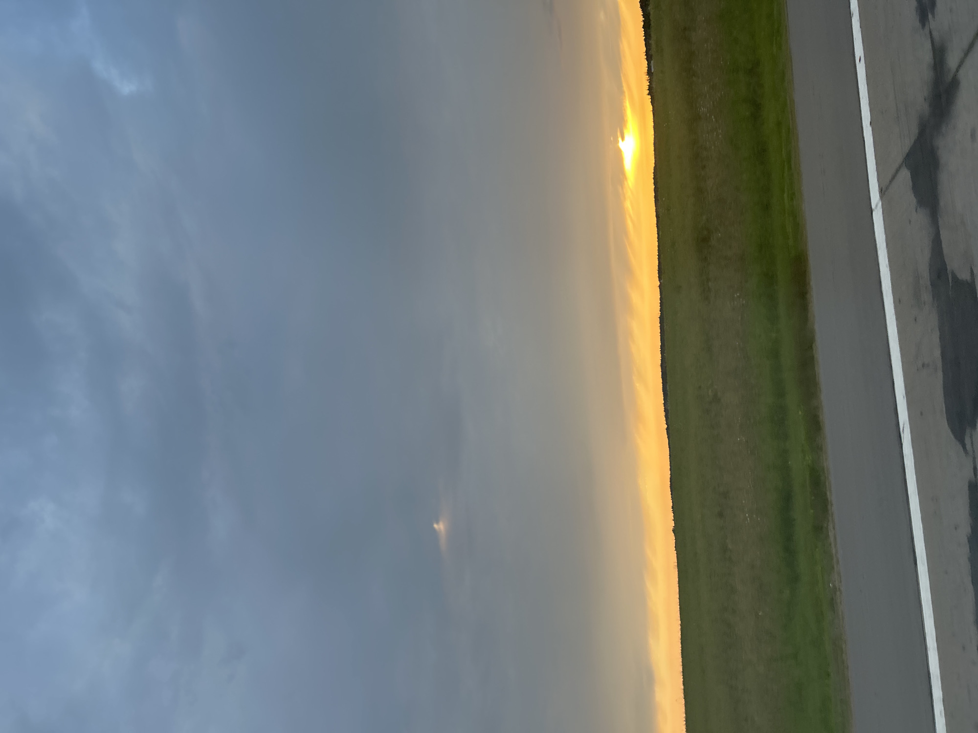 The image depicts a rural landscape in the vicinity of Ozyaritska-Slabadski rural council, Belarus. The scene is characterized by a wide expanse of green grassland under a dramatic sky. The sun is setting, casting a warm, golden glow on the horizon and illuminating the clouds from below. The sky transitions from a bright, orange-yellow band near the horizon to a darker, overcast gray overhead, with a few wispy clouds scattered throughout. In the distance, a dark line of trees marks the edge of the field. In the foreground, a section of asphalt road with a white line is visible, suggesting it is a road or path bordering the field. There are no people or animals present in the image. The overall atmosphere is one of natural beauty and tranquility, with the sunset creating a striking visual contrast.