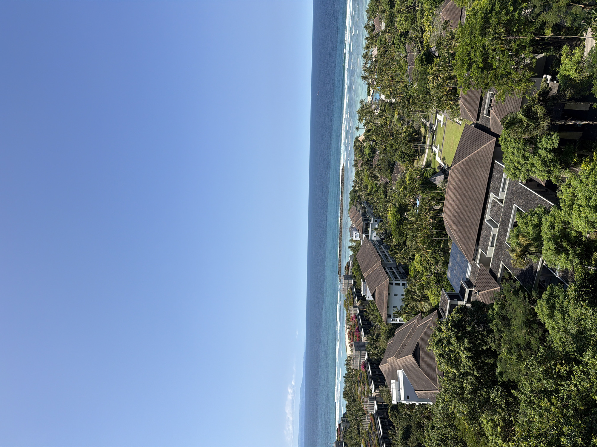 The image shows an aerial view of the coastline in Kuta Selatan, Indonesia, with a clear blue sky dominating the left side. The ocean is visible, with waves breaking along the shoreline. Buildings with brown roofs are interspersed with lush green trees. Palm trees are abundant, suggesting a tropical environment. Some buildings appear to have modern architectural designs.