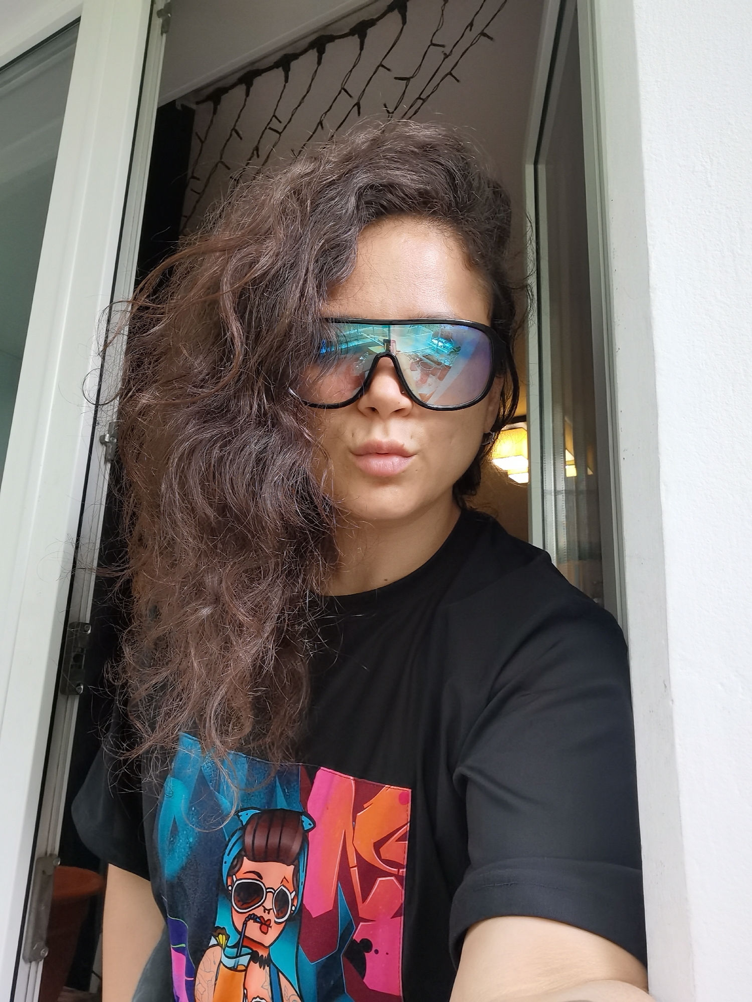 A close-up portrait shows a person with long, voluminous, dark brown, curly hair, posing for the camera with pouty lips. They are wearing large, reflective, blue-tinted, shield-style sunglasses that obscure their eyes. The person is dressed in a black t-shirt featuring a vibrant, graffiti-style graphic on the front, depicting a stylized female character with sunglasses, holding a drink with a straw, set against colorful, abstract letters.
The person appears to be leaning into a doorway or window frame, with a white frame visible on the left and a white wall on the right. Through the opening behind them, a darker interior space is visible, with decorative string lights strung horizontally across the top of the frame. The reflections in the sunglasses reveal an inverted image of an indoor scene, including what appears to be a ceiling or light fixture and other interior details, suggesting the photo was taken indoors. The lighting indicates it is daytime. The overall mood conveyed is confident and playful. The scene is located in Chaikovsky, Russia.