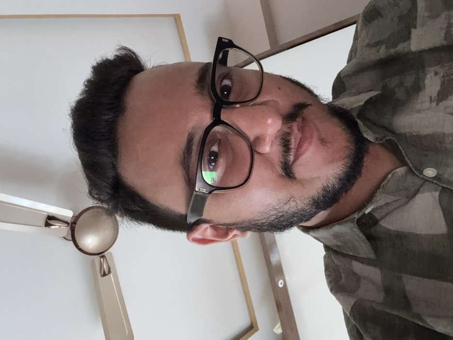 A close-up, upward-angled portrait features an adult male. He has dark hair, a neatly trimmed beard, and a mustache. The subject wears dark-rimmed rectangular eyeglasses. His eyes are directed at the viewer, and his mouth shows a slight, closed-lip smile. He is dressed in a collared button-up shirt with a patterned design, predominantly in shades of olive green and light beige. The background is an interior setting with a white ceiling. A cream-colored three-bladed ceiling fan is partially visible in the upper left portion of the frame. Decorative trim, possibly wood, outlines a recessed area on the ceiling. The image provides no specific visual indicators to confirm a location within Howrah, India.