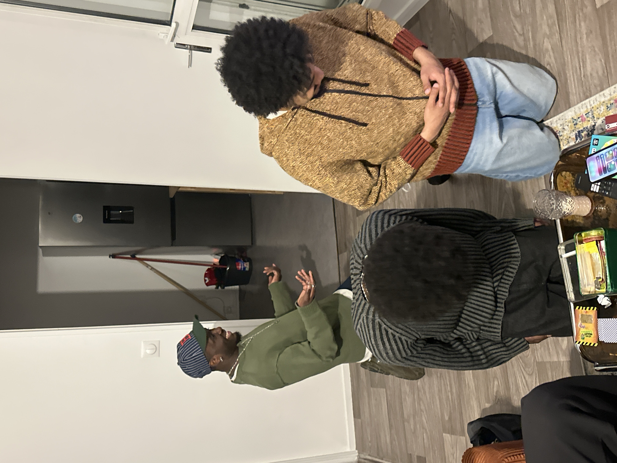 Three individuals are gathered indoors around a glass-topped table. One person wears a patterned brown and red hooded sweatshirt and light-wash jeans, kneeling beside the table. Another is wearing a striped grey and black hooded sweater, also kneeling at the table. A third individual, wearing a green hoodie and a striped hat, is seated on the floor. Items on the table include an iPhone, remote control, glass ornament, playing cards, and a container of snack food. The floor is light-colored wood laminate. A stainless steel refrigerator is visible through an open doorway. This appears to be a casual social gathering in an apartment. There are no indications that this location is in Paris, France.
