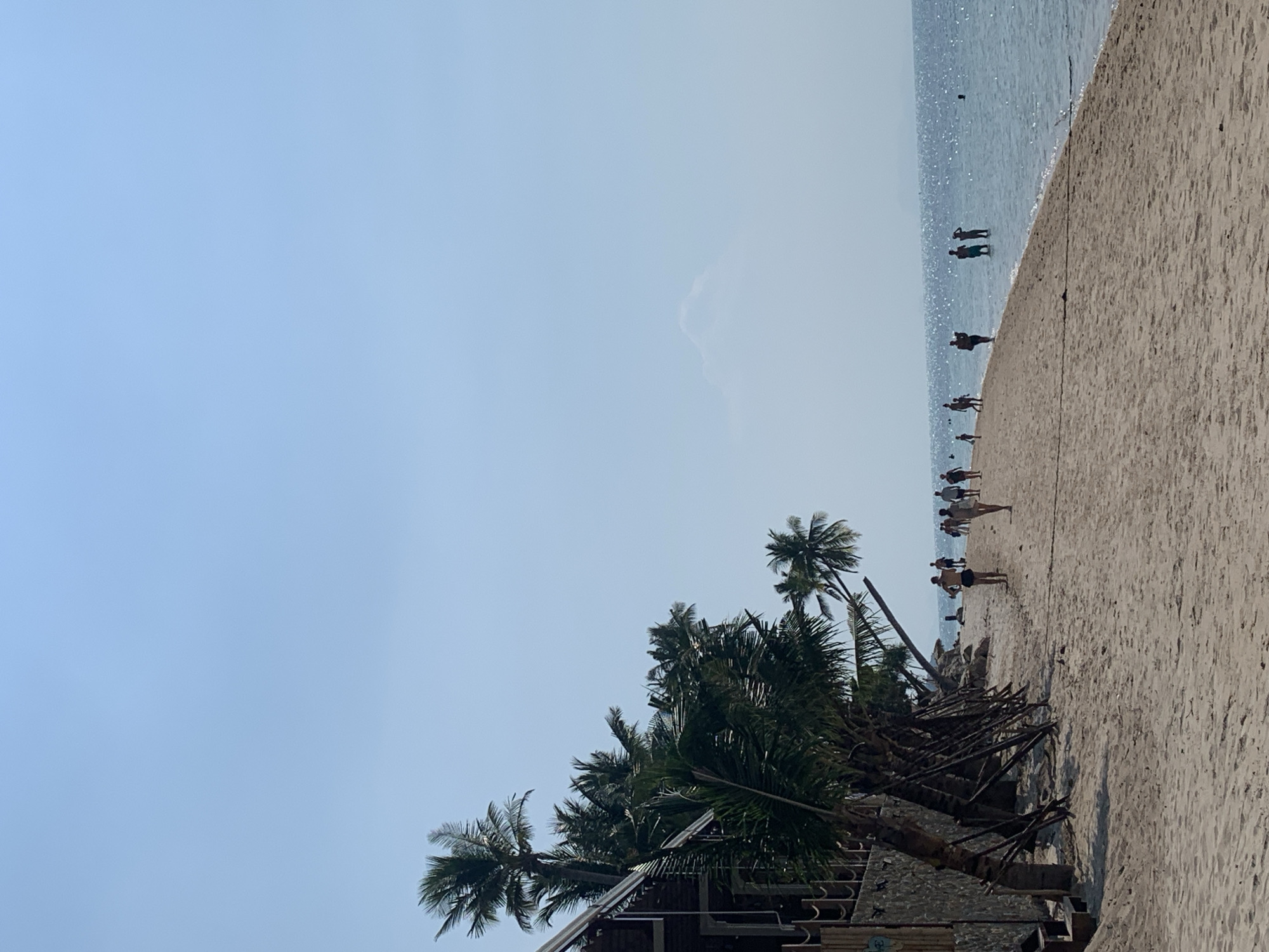 The image depicts a beach scene in Baan Tai, Thailand. The sky is a pale blue, transitioning to the slightly darker blue of the sea. The sandy beach stretches from the right foreground, extending toward the horizon. A line of people are standing and walking along the beach near the water’s edge. Two individuals are standing in the water.
In the lower left corner, there are palm trees and part of a structure, likely a building or shelter. The palm trees are leaning towards the right. Fallen palm fronds are visible in front of the trees. The overall impression is of a tropical beach scene.