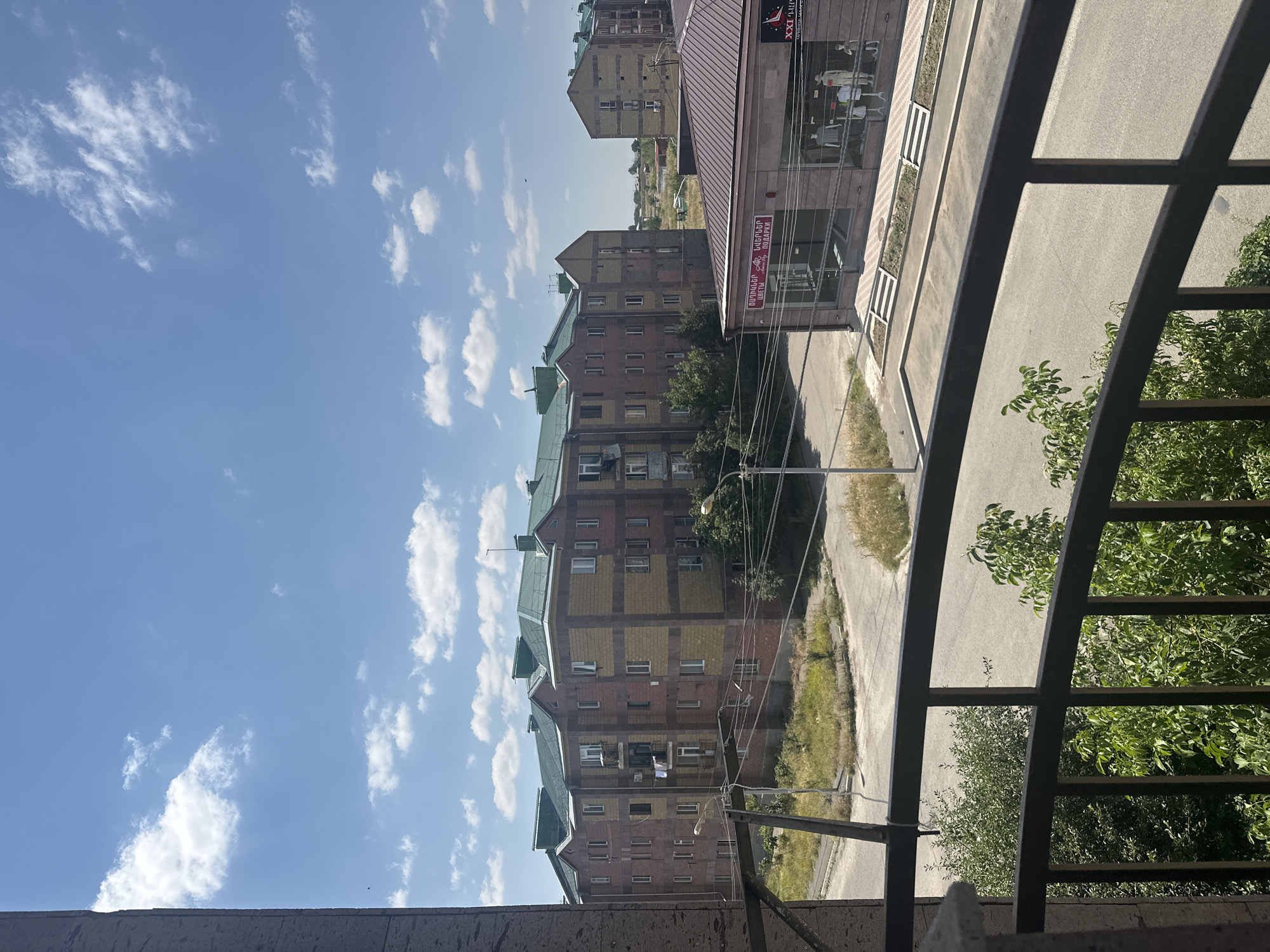 The image depicts a sunny day in Gyumri, Armenia. In the foreground, a dark metal balcony railing partially obstructs the view of lush green trees. Beyond the trees, a paved road runs horizontally across the frame. To the right of the road, a low-rise building with a dark brown roof and a stone facade features large display windows showcasing mannequins dressed in clothing. Above the windows, text in Armenian characters is visible. Further in the background, a large apartment building with a green roof, constructed with brick and yellow facade elements, dominates the scene. Several smaller buildings and scattered greenery are visible in the distance, under a bright blue sky dotted with white clouds.