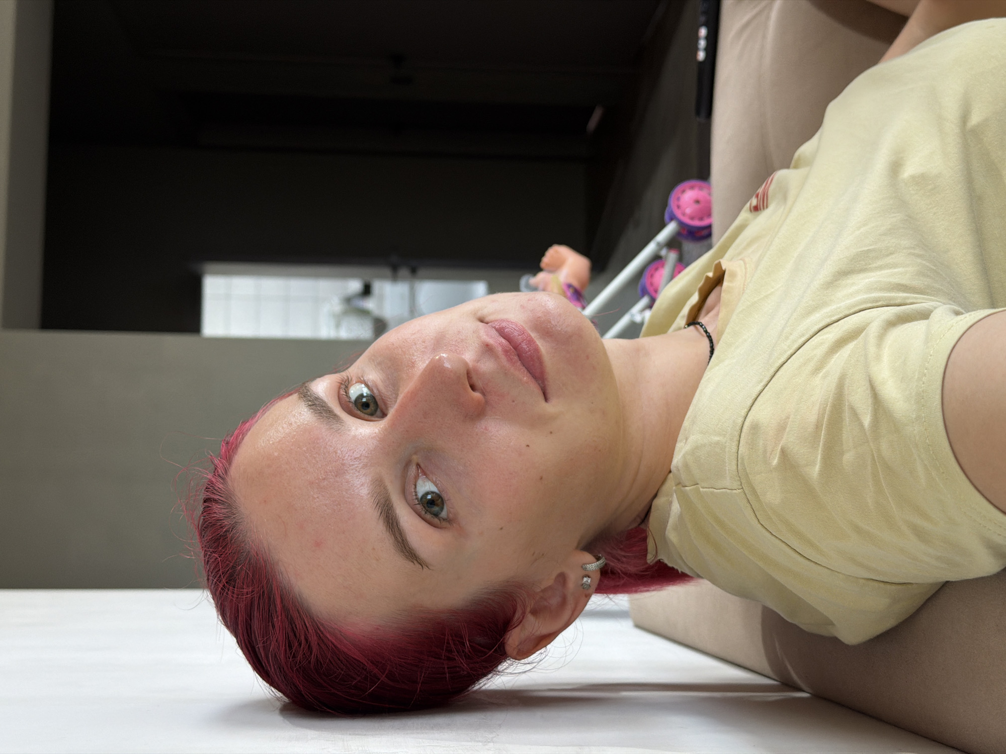 The image shows a young woman with bright pink hair leaning against a white wall. She is wearing a light yellow t-shirt and a black beaded necklace. Her expression is a gentle smile as she looks towards the camera. Her eyes are green and her skin has a slight flush. In the background, a doorway leads to a darker room, and to the right, a small pink and purple toy stroller is visible. The woman is seated on a light brown sofa. The overall setting appears to be indoors, likely in a residential apartment. There are no visible indications connecting this image to the city of Chaikovsky, Russia.