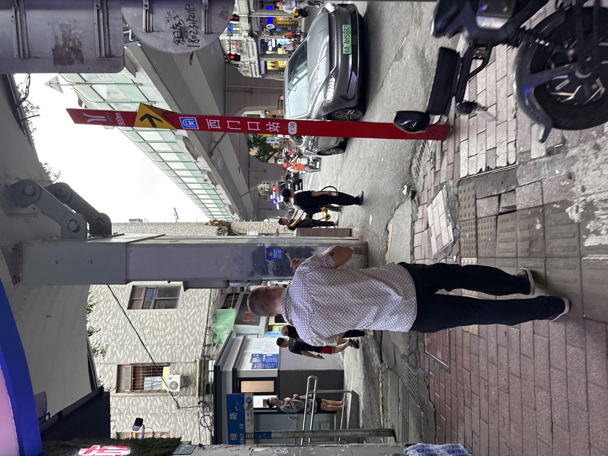 The image depicts a street scene in Yuexiu District, China. In the foreground, a man with short hair, wearing a white patterned short-sleeved shirt and black trousers, is walking away from the camera. He is on a sidewalk paved with red and grey bricks. To his left, a blue sign with Chinese characters and the text "ANLV LU" is visible on a white structure. Further down the sidewalk, a man in a grey shirt and shorts stands facing a building entrance with metal railings. Behind them, other individuals are walking, including a person in a black t-shirt and red shorts.
The background shows a street with vehicles, including a grey car with a license plate starting with "AH5882," a police car with flashing lights, a scooter, and several other motorcycles and bicycles. An elevated walkway with a green railing crosses overhead. A red pole with signage, including a directional arrow and Chinese characters, is positioned near the street. Traffic lights are visible. Buildings line the street, with windows and an air conditioning unit on one facade. The sky is overcast.