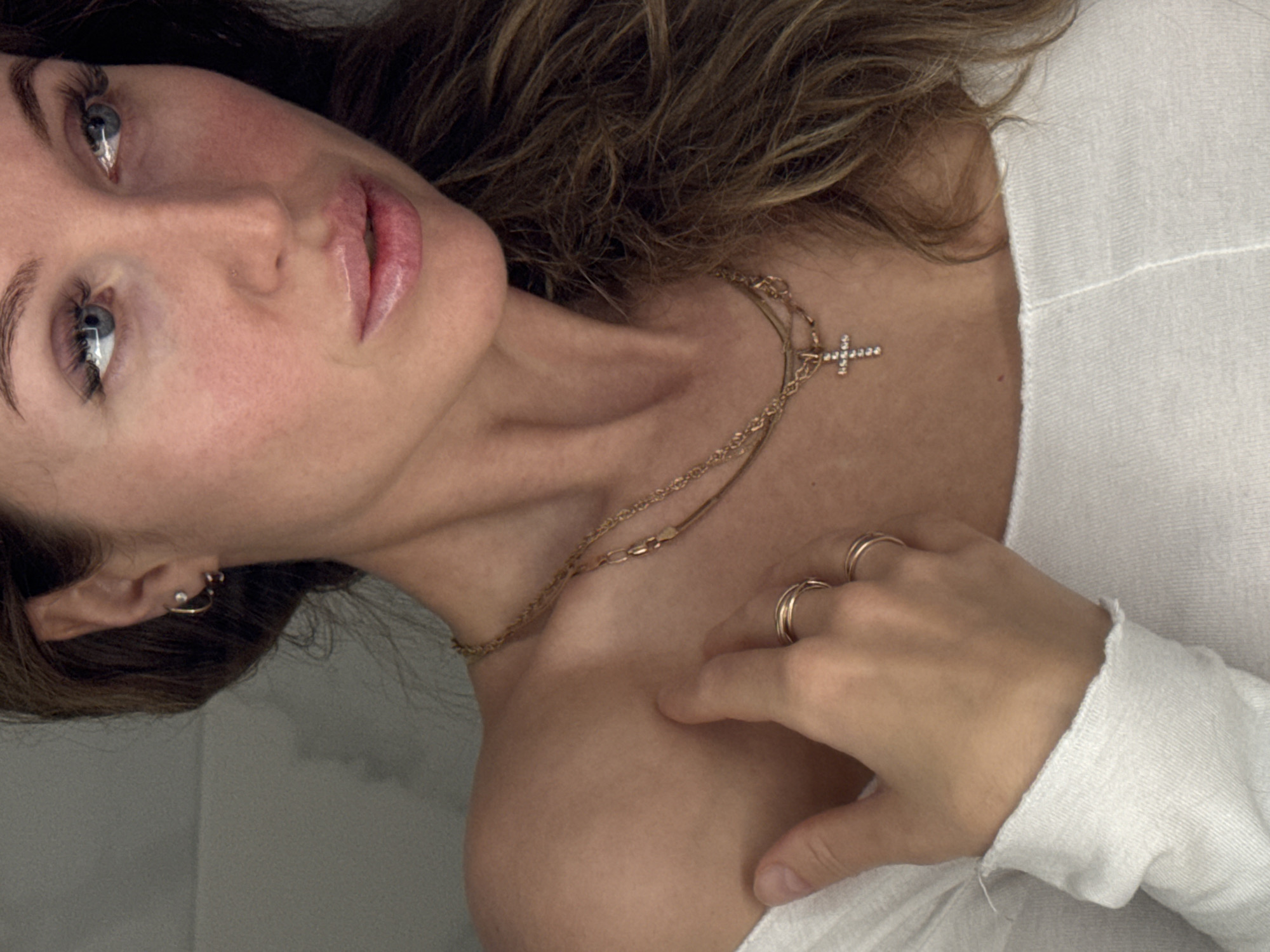 A woman is pictured from a close-up, slightly angled perspective. Her gaze is directed upwards and towards the left. She has fair skin, light eyes, and her lips appear to have a glossy finish. Her hair is light brown and partially visible. She is wearing a white, long-sleeved top. Around her neck is a gold necklace with a cross pendant. On her hand, which rests on her shoulder, she is wearing two gold rings. A small earring is visible in her ear. The background appears to be a plain, light-colored wall. There is no evidence to suggest the location is Chaikovsky, Russia.