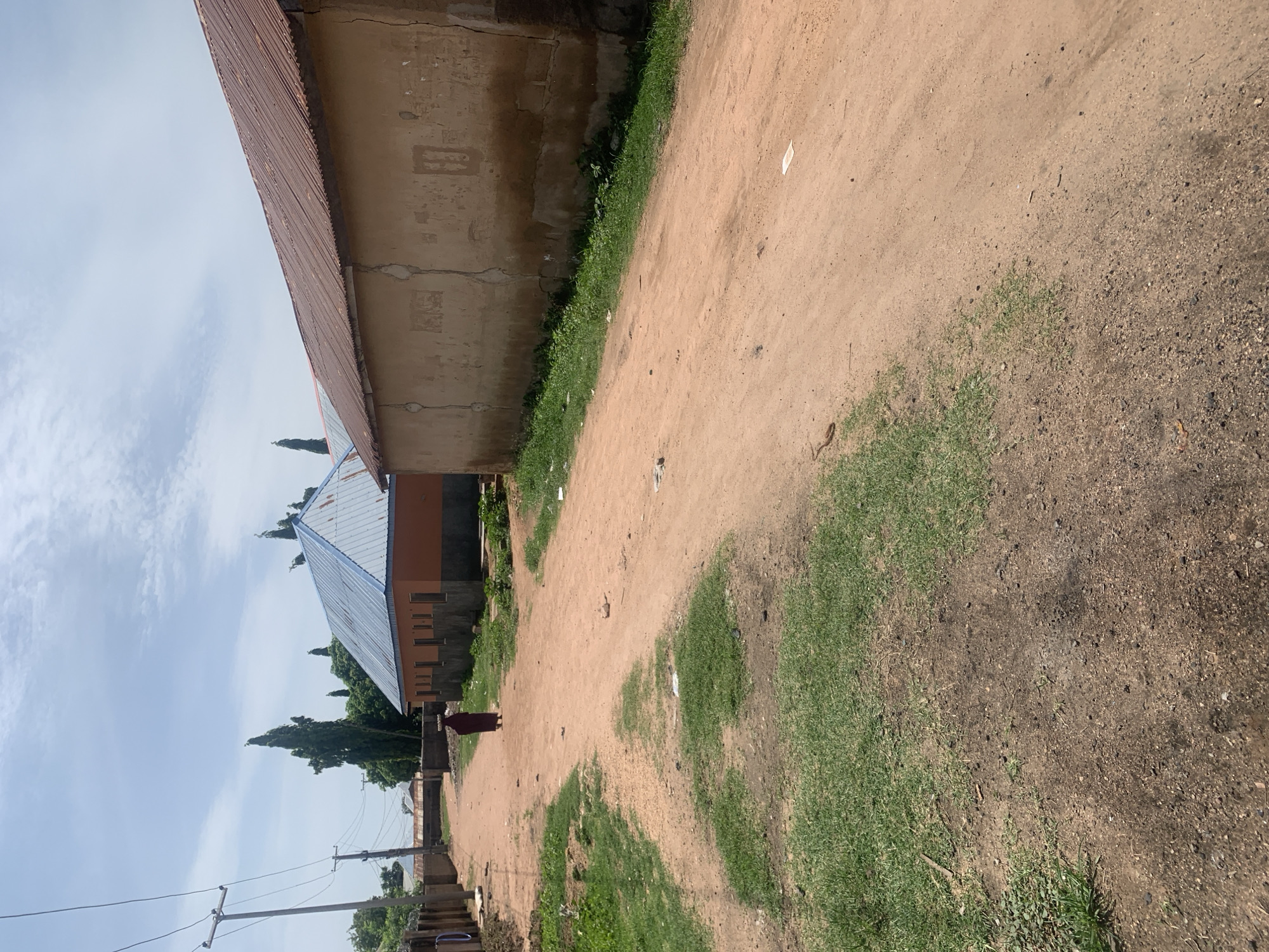 In Jauro Yinu, Nigeria, a dirt road runs through a residential area, flanked by buildings. On the right, a long, single-story building with a rusted corrugated metal roof has a rough, light brown textured wall. On the left, another building with an orange facade and a blue corrugated metal roof is visible. A person wearing a dark red garment walks away from the camera down the dirt road, carrying a load on their head. The ground is a mix of red dirt and patches of green grass. Power lines run diagonally across the sky, supported by wooden utility poles. Tall, thin trees are scattered throughout the scene. The sky is cloudy with patches of blue.