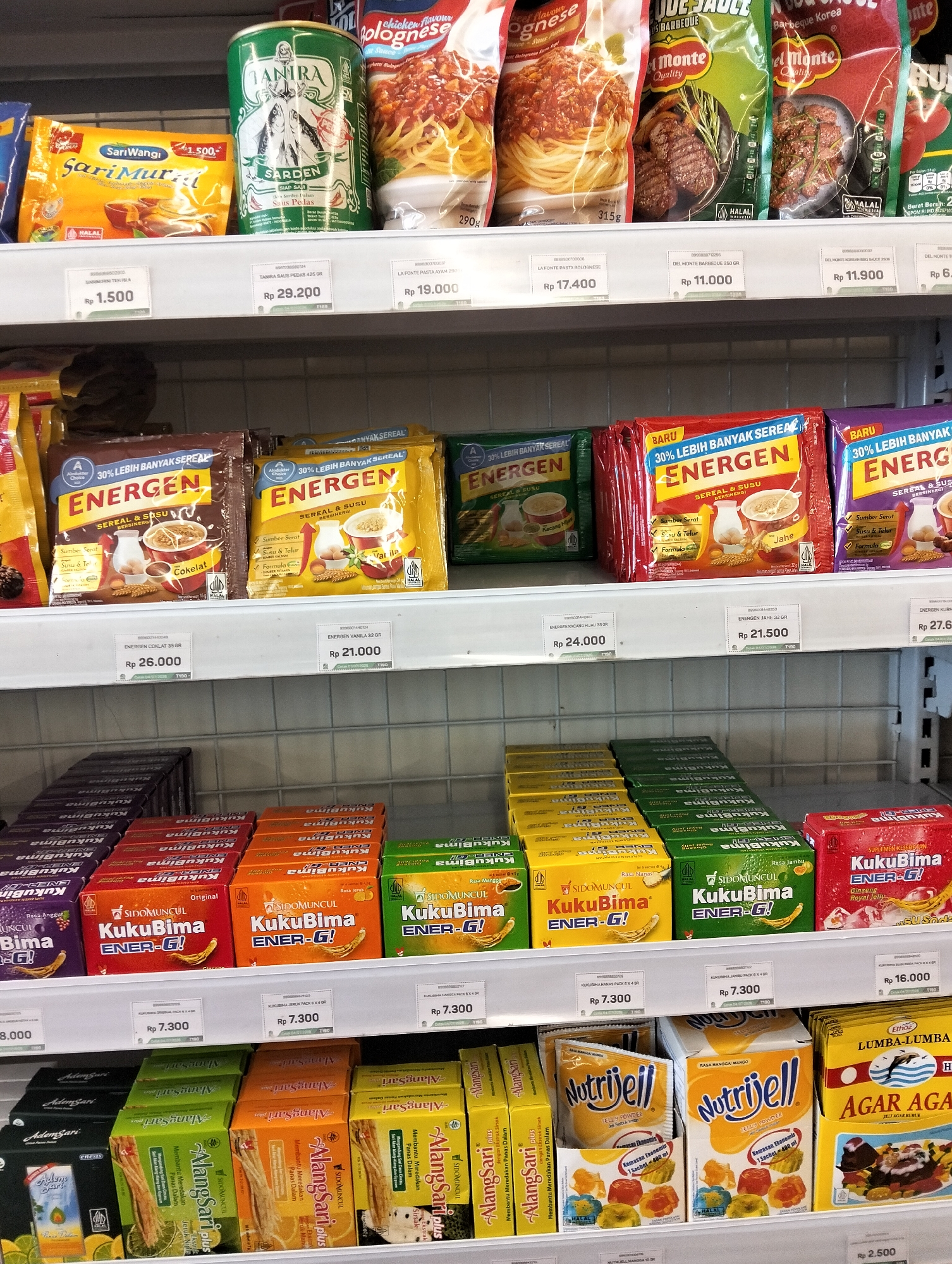 The image displays a retail shelf in a store located in Muncar, Indonesia, filled with various packaged food and beverage products. The shelves are made of white metal grating. On the top shelf, there are cans of "SariWangi Sarden" and packs of "Energen" cereal and drinks in different flavors like chocolate, vanilla, kacang hijau (mung bean), and jahe (ginger). Below that, the second shelf contains more "Energen" packs, and then packs of "KukuBima Ener-G" in various flavors such as original, rasa nanas (pineapple), and rasa jambu (guava). The bottom shelf showcases "AdemSari Plus" and "Nutrijell" jelly powder in mango flavor, along with "Agar Agar" gelatin. The products are neatly arranged, with price tags visible underneath some of them. The lighting appears to be from overhead store lights, illuminating the products. There are no people visible in the image.