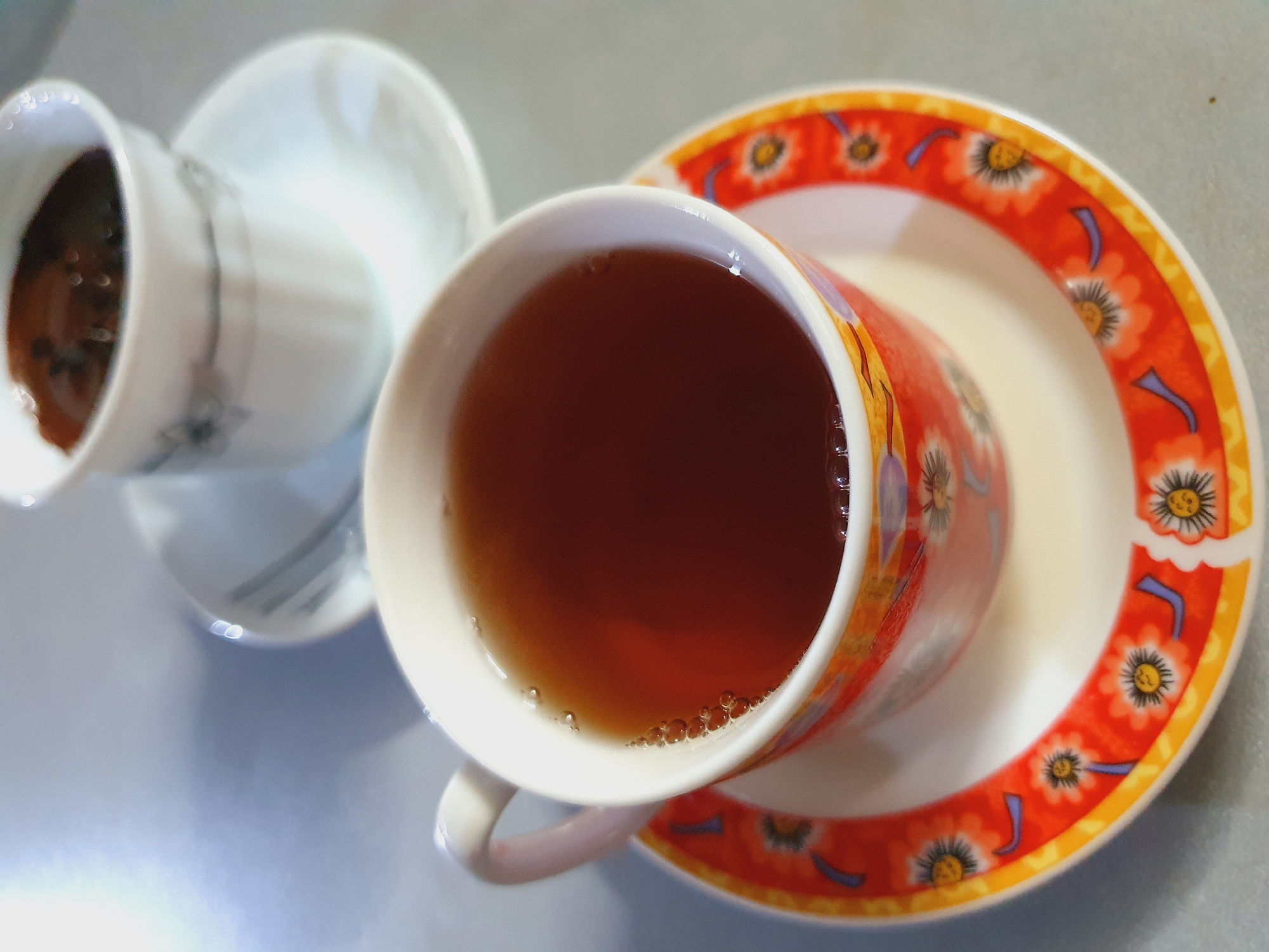 A close-up, overhead shot shows two teacups with saucers on a light gray surface, possibly a table. The cup in the foreground is a white ceramic teacup with a brightly colored floral pattern in shades of orange, yellow, and red around its base. It is filled with a dark amber liquid, likely tea, with small bubbles visible on the surface. The saucer beneath it mirrors the floral design of the cup. Behind this, slightly out of focus, is another white teacup with a delicate gray floral pattern, containing a dark brown liquid, suggesting coffee. Its white saucer is also visible. The lighting suggests an indoor setting, possibly a cafe or home environment in Shiraz, Iran.