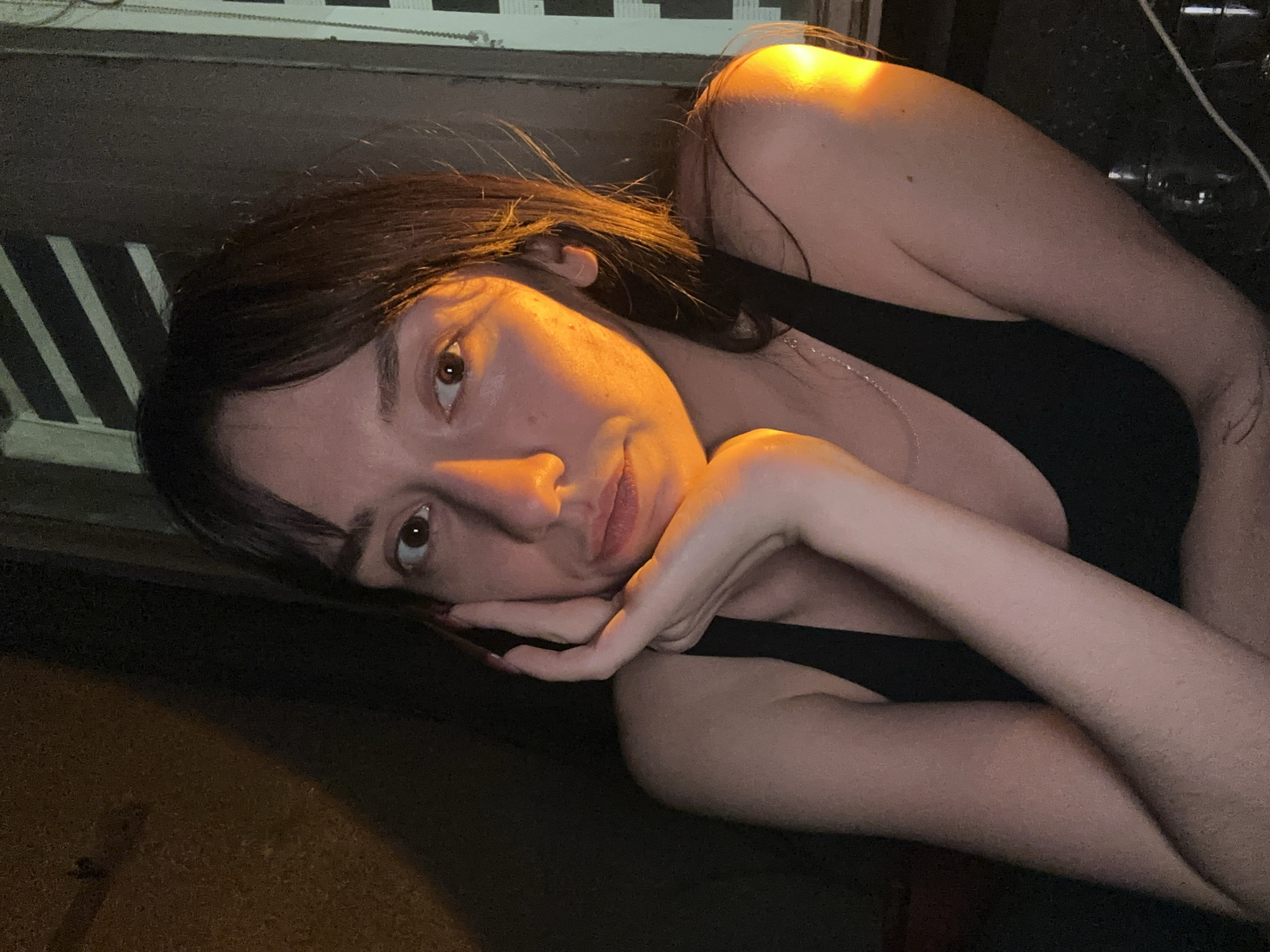 A young woman lies with her head resting on her hand. She has dark hair with bangs. She wears a black tank top and a silver necklace. The lighting is warm. The location is unknown.