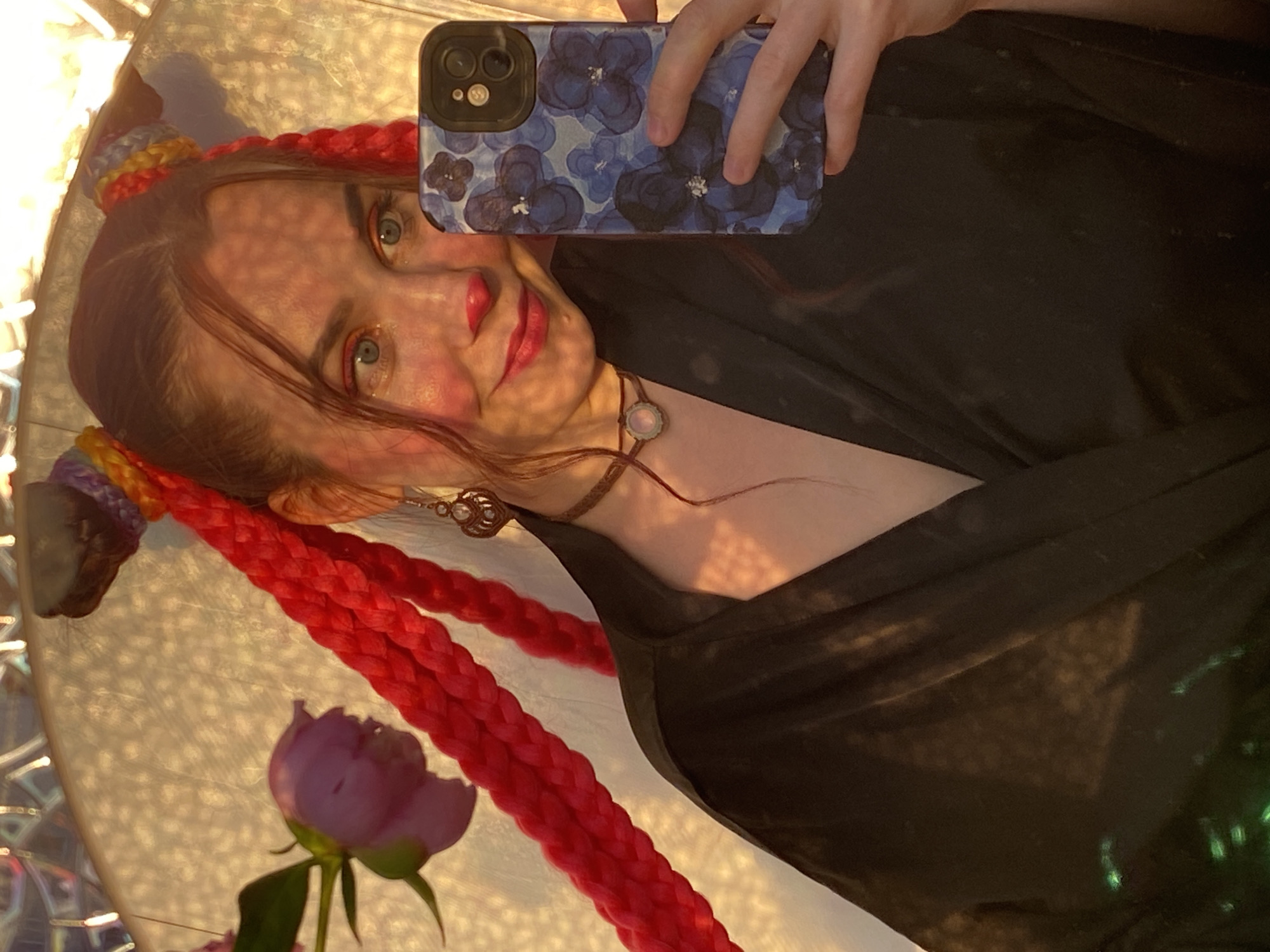 This is a medium shot, selfie-style photograph of a young woman with bright red braided pigtails that are tied up in two buns on top of her head with colorful accents. She is wearing a black garment, possibly a robe or kimono, and a choker necklace with a pale pink pendant. She has blush on her cheeks, red lipstick, and a red dot on her nose, giving her a clown or character-like appearance. Her eyes are open and she is looking towards the camera. In the background, there is a blurred, light-colored surface with some texture and a large, indistinct light fixture or decoration. To the left of her head, there is a partially visible pink flower. The image appears to be taken indoors, possibly in a home or studio setting. The location in the city of Chaikovsky, Russia, cannot be determined from the visual information present in the image.