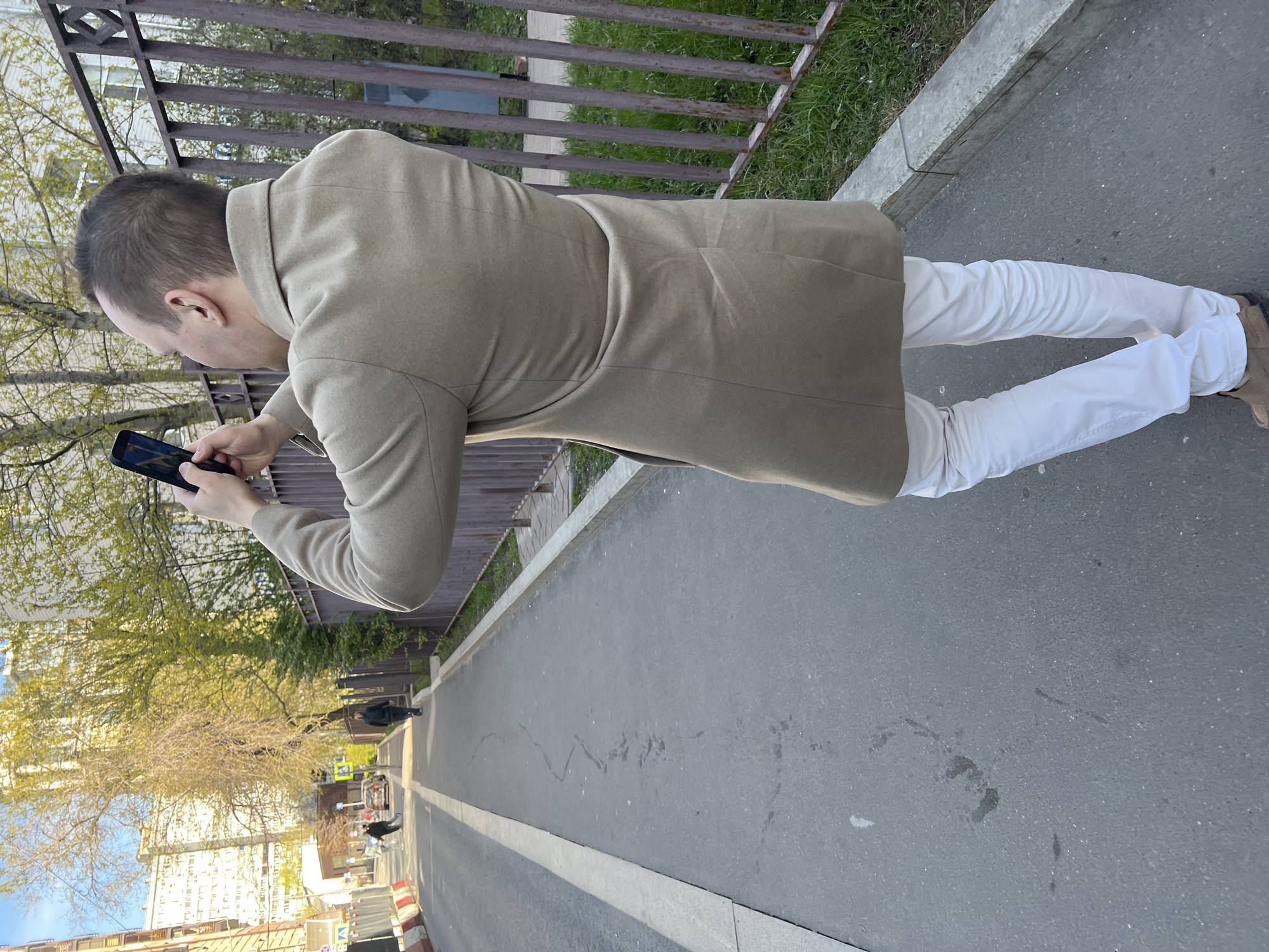 A man with short hair, wearing a tan coat and white pants, stands on a gray asphalt road, holding a phone. The road has a white painted line marking its edge. A low metal fence runs along the side of the road, separating it from a grassy area. In the background, there are buildings and trees, consistent with an urban environment. Based on the style of buildings visible, and the mention of "Moscow, Russia," the setting is likely a residential area in Moscow.