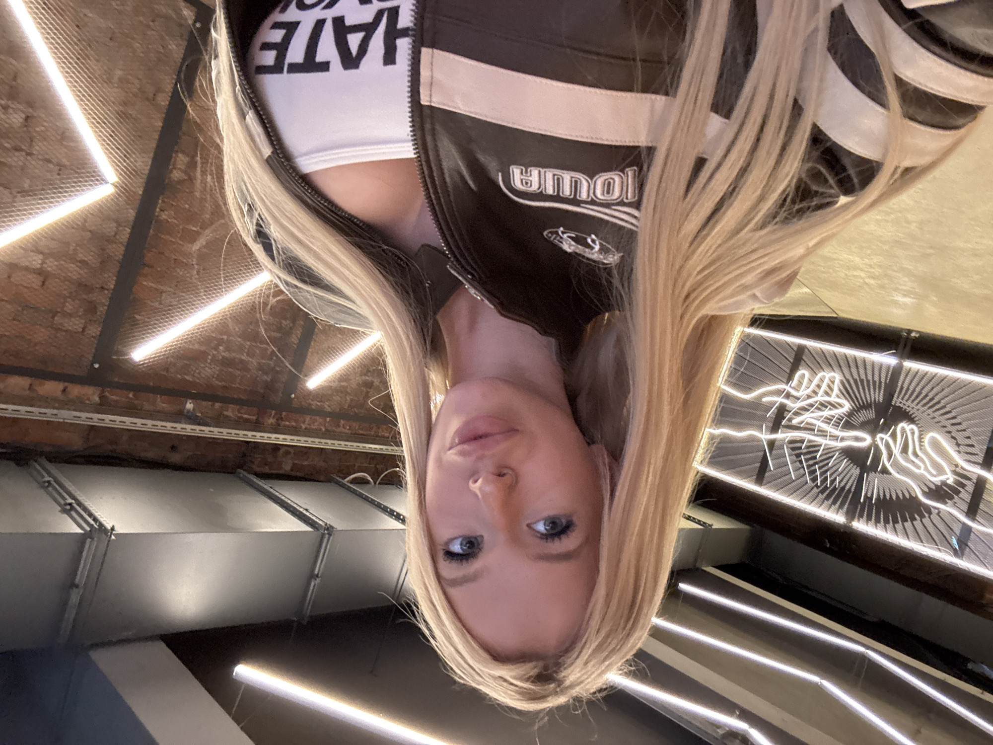 This is a selfie of a young woman with blonde hair. She is wearing a white crop top with the word "HATE" printed in large black letters across the chest. Over the crop top, she wears a brown and white racing-style jacket with the word "IOWA" written in white lettering on the left chest. She has blue eyes and is looking directly at the camera. The background is blurry, but appears to be an indoor industrial-style space with exposed brick walls and ductwork. There are also bright neon lights that create geometric patterns on the ceiling. The overall lighting suggests it is nighttime or indoors with artificial lighting. The image is oriented upside down. The location context provided is Saint Petersburg, Russia.