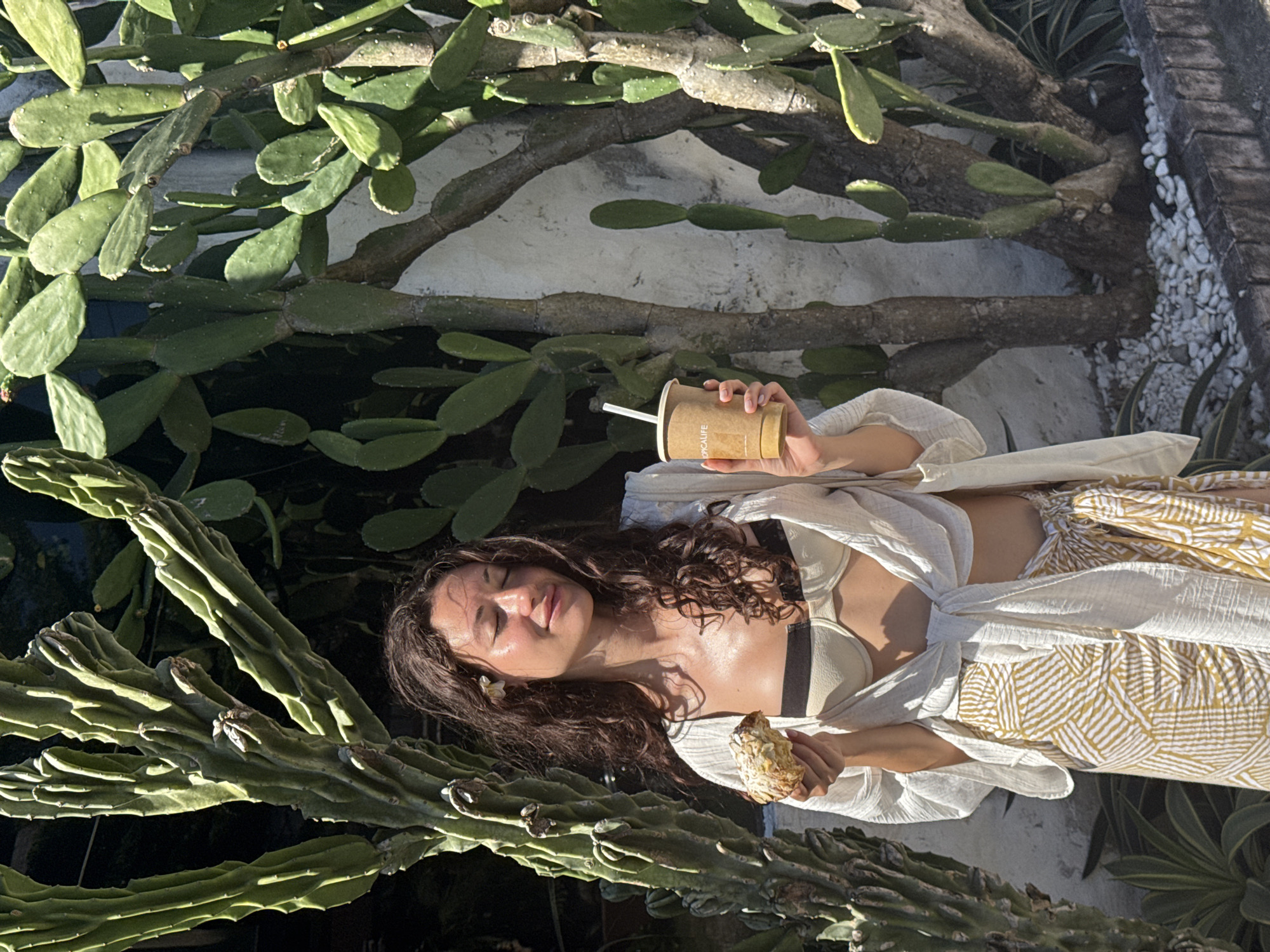 A light-skinned woman with long, wavy brown hair is sitting outdoors amidst cacti, holding a paper cup with a straw in her right hand and a pastry in her left. She is wearing a light-colored bikini top, a white blouse, and a tan patterned wrap around her waist. A small white flower is tucked into her hair near her left ear. The background includes various types of cacti, a white wall, white rocks and other plants. The location is the city of North Kuta, Indonesia.