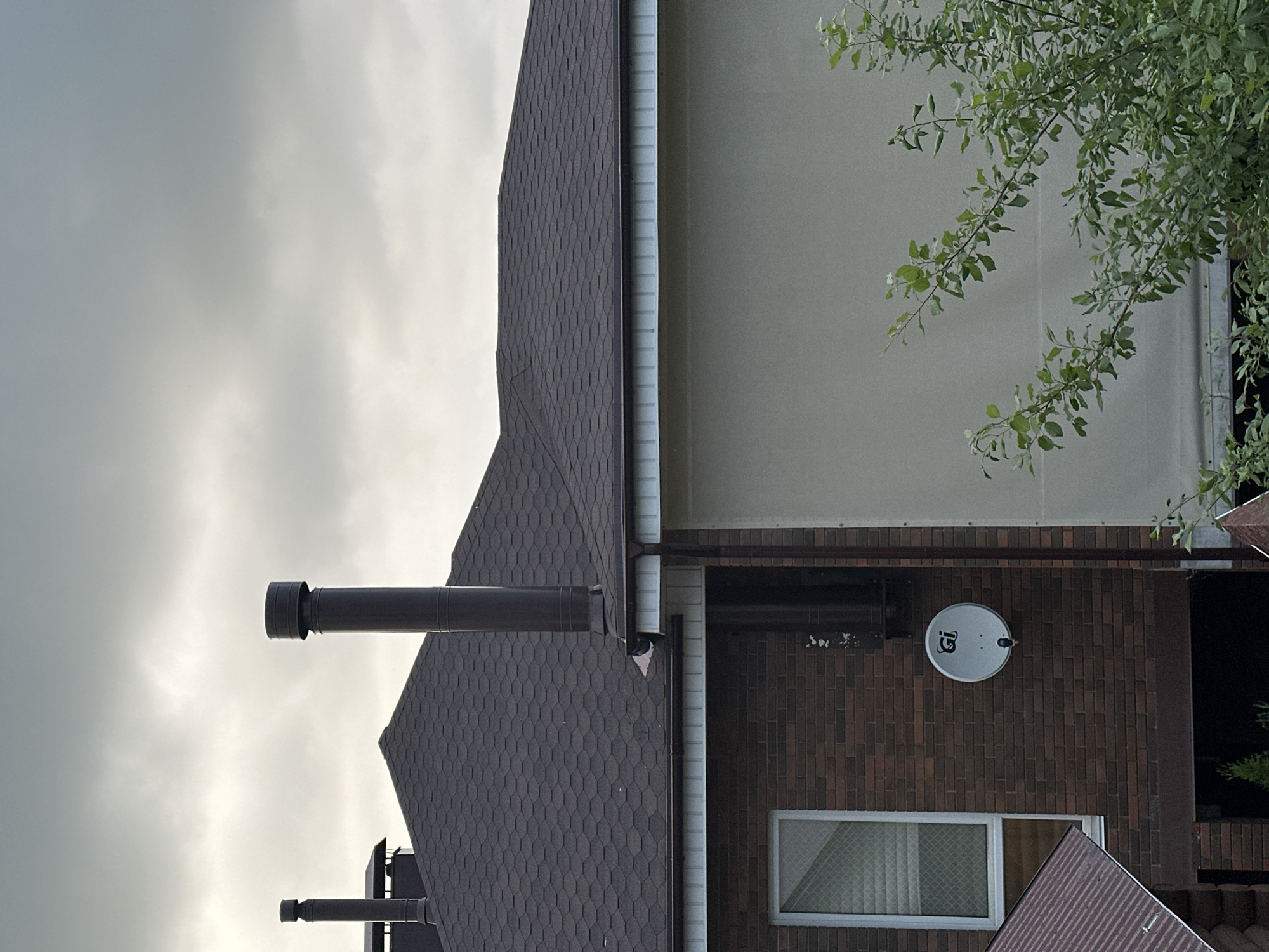 The image shows the upper portion of two adjacent buildings. The building on the left has a dark brown brick facade and a dark brown asphalt shingle roof. A white window with a diamond pattern is visible on the brick facade. Two dark brown chimneys extend from the roof of this building. A satellite dish, white with a "GI" logo, is mounted on the brick wall. The building on the right has a light beige, textured exterior wall and a dark brown asphalt shingle roof. A large, light-colored retractable awning or screen covers a significant portion of the right building's facade. Green foliage from a tree is visible in the lower right corner of the image. The sky is overcast with gray clouds. There are no people or animals visible in the image. The scene appears to be a residential area, likely suburban, given the architectural style and presence of residential amenities like chimneys and a satellite dish. There are no specific landmarks or indications within the image that would allow for pinpointing a location within Nizhny Novgorod, Russia.