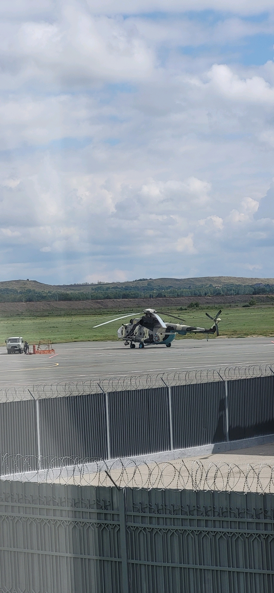 An outdoor, medium-shot captures a camouflage military helicopter situated on an airport tarmac. The helicopter's blades are slightly blurred, suggesting potential movement or a long exposure. To the left of the helicopter, a white, four-door utility vehicle is parked, with two individuals standing beside it. Between the vehicle and the helicopter, an orange barrier is visible. The tarmac extends into the background, where a grassy field with scattered trees and rolling hills forms the landscape. The sky is predominantly filled with white, fluffy clouds against a light blue backdrop. In the foreground, a grey metal fence with barbed wire atop it partially obscures the view of the tarmac and the distant scene. The scene appears to be an operational airfield. The specific location in the city of Oskemen, Kazakhstan, is not identifiable from the image content.