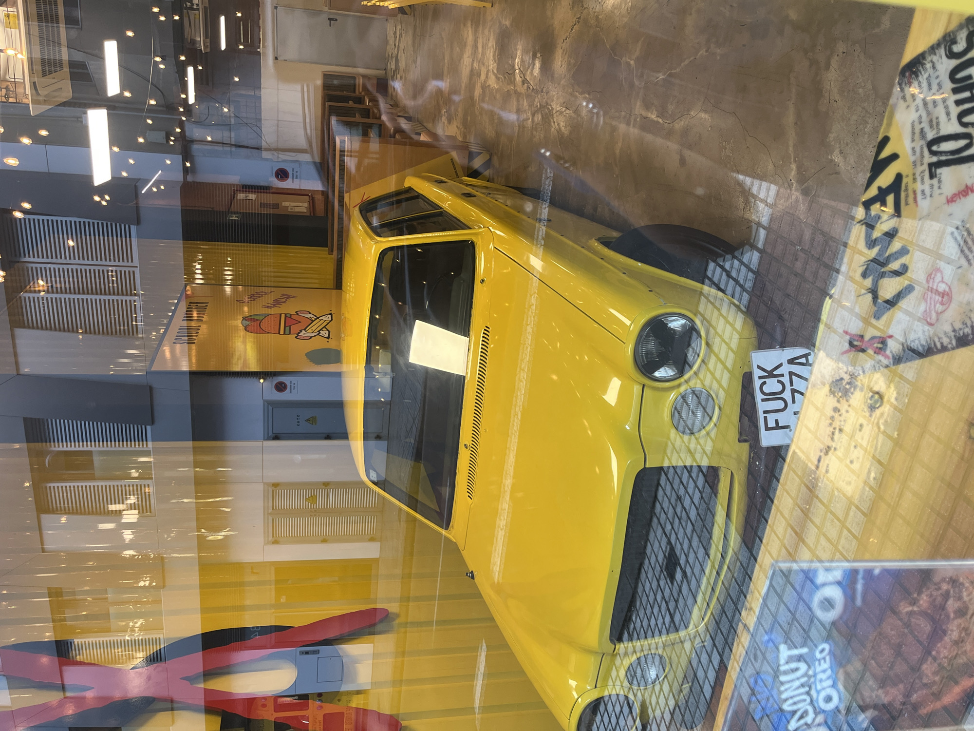 The image displays a vibrant yellow, vintage-style compact car, likely positioned as an exhibit inside a shop window in Las Palmas, Spain. The car, with its round headlights and black grille, prominently features a white license plate that reads "FUCK 77A". The photograph is taken through a highly reflective glass window, superimposing external elements onto the internal scene.
Reflections of bright, vertical and scattered circular light fixtures, along with outlines of buildings and possibly the sky, are visible across the entire image. Inside the shop, behind the yellow car, a yellow wall is adorned with a large graphic of a rocket-like pencil character and the words "SCHOOL BURGER," suggesting the establishment might be a burger joint. The interior floor is a textured concrete.
Further visible text includes "DONUT OREO" on a display item in the foreground, reinforcing the idea of a food-related business. Other partially legible text from reflected signs or posters includes "ketch," "TABW I," "driw," and fragments like "100toa Inosit," "sagraud," and "ERRAR." The bright lighting conditions suggest it is daytime, with both natural light and interior artificial lighting illuminating the scene. The overall impression is a quirky and eye-catching commercial display.
